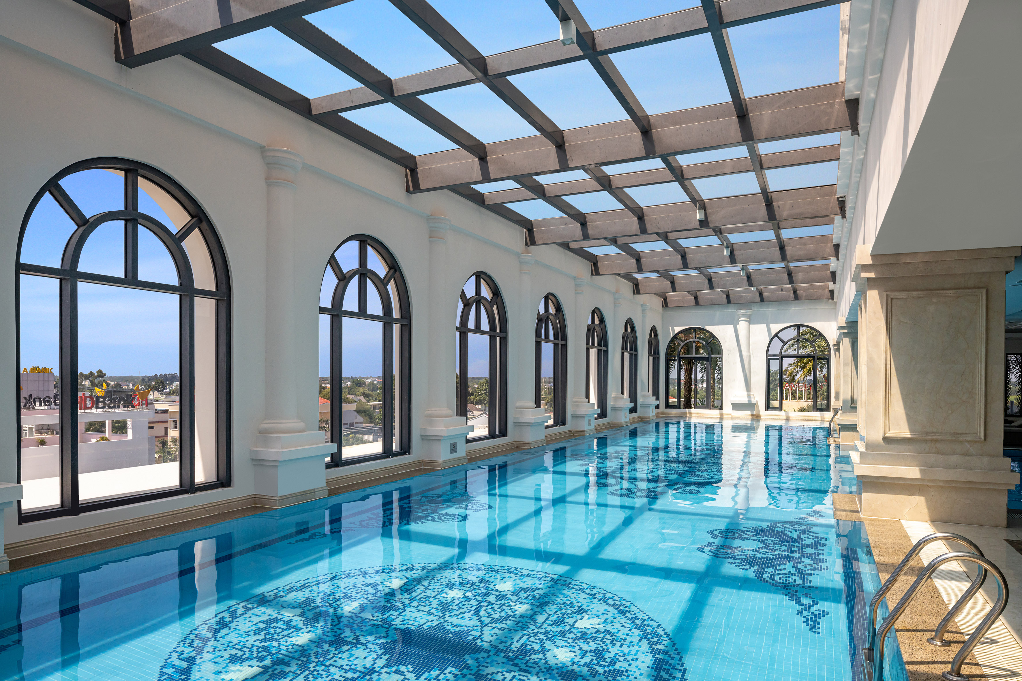Hotel indoor swimming pool with mosaic tiles, arched windows, and glass skylight.