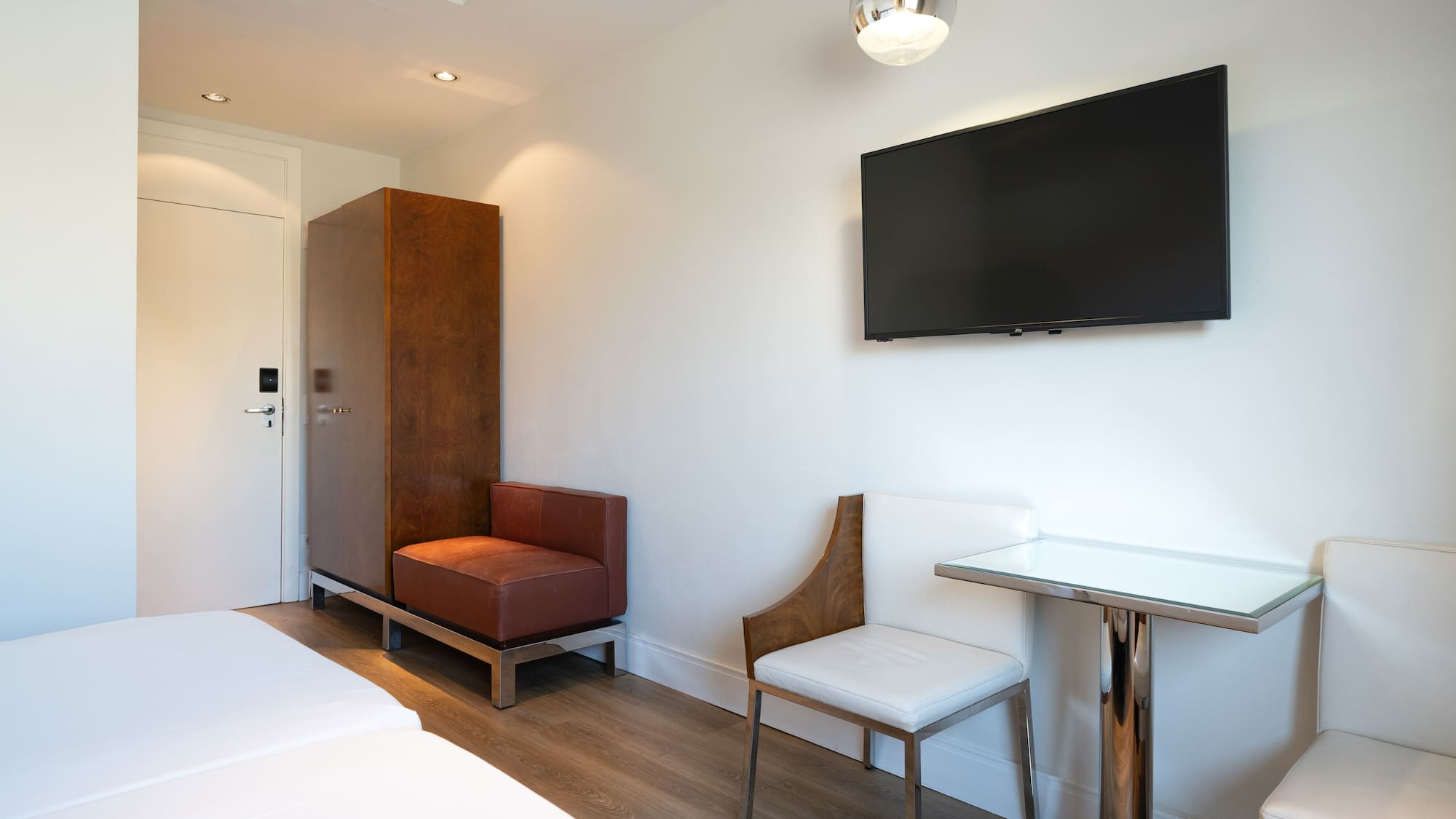Modern hotel room with wall TV, wood wardrobe, bed, table, and chairs.