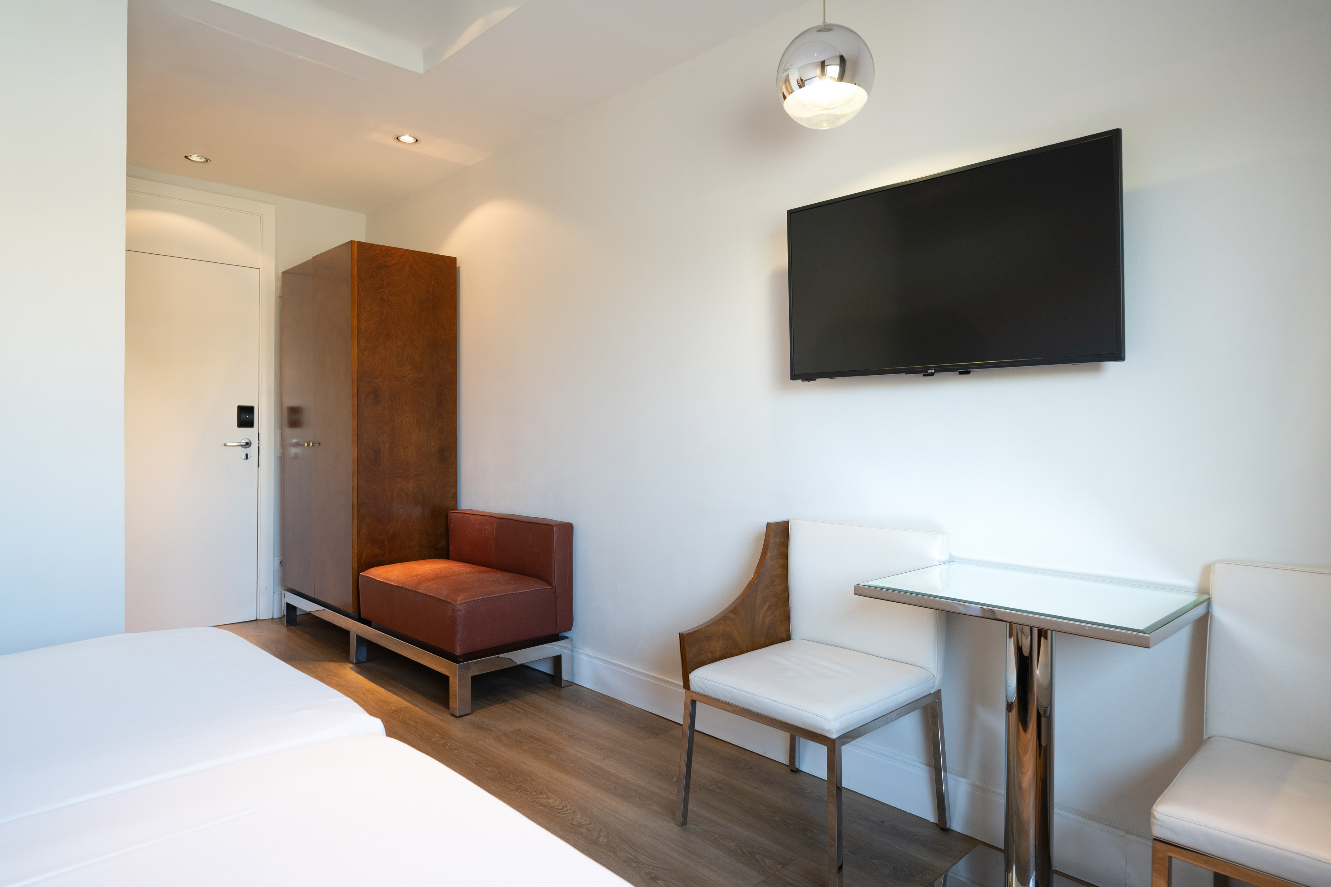 Modern hotel room with wall TV, wood wardrobe, bed, table, and chairs.