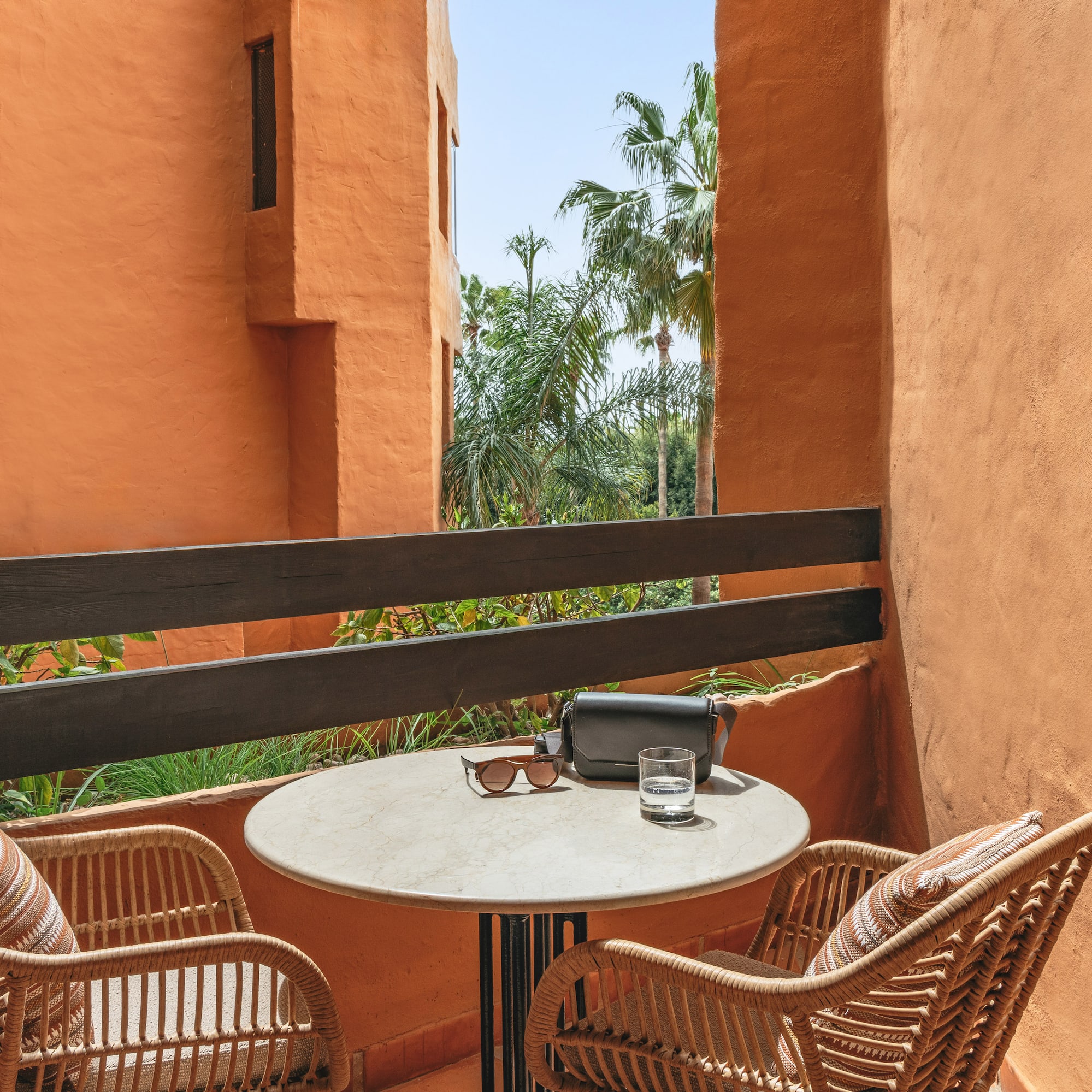 Terracotta balcony with wicker chairs, table, sunglasses, and palm trees.