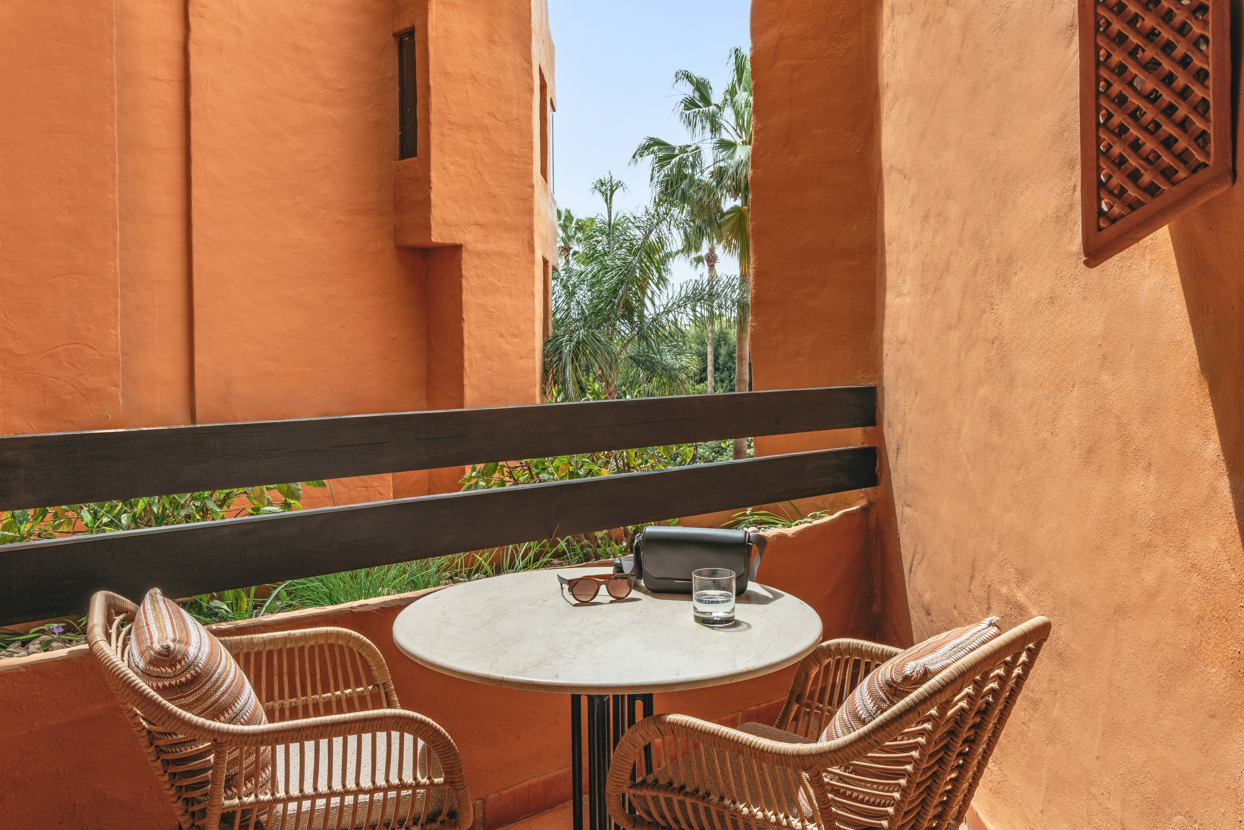 Terracotta balcony with wicker chairs, table, sunglasses, and palm trees.