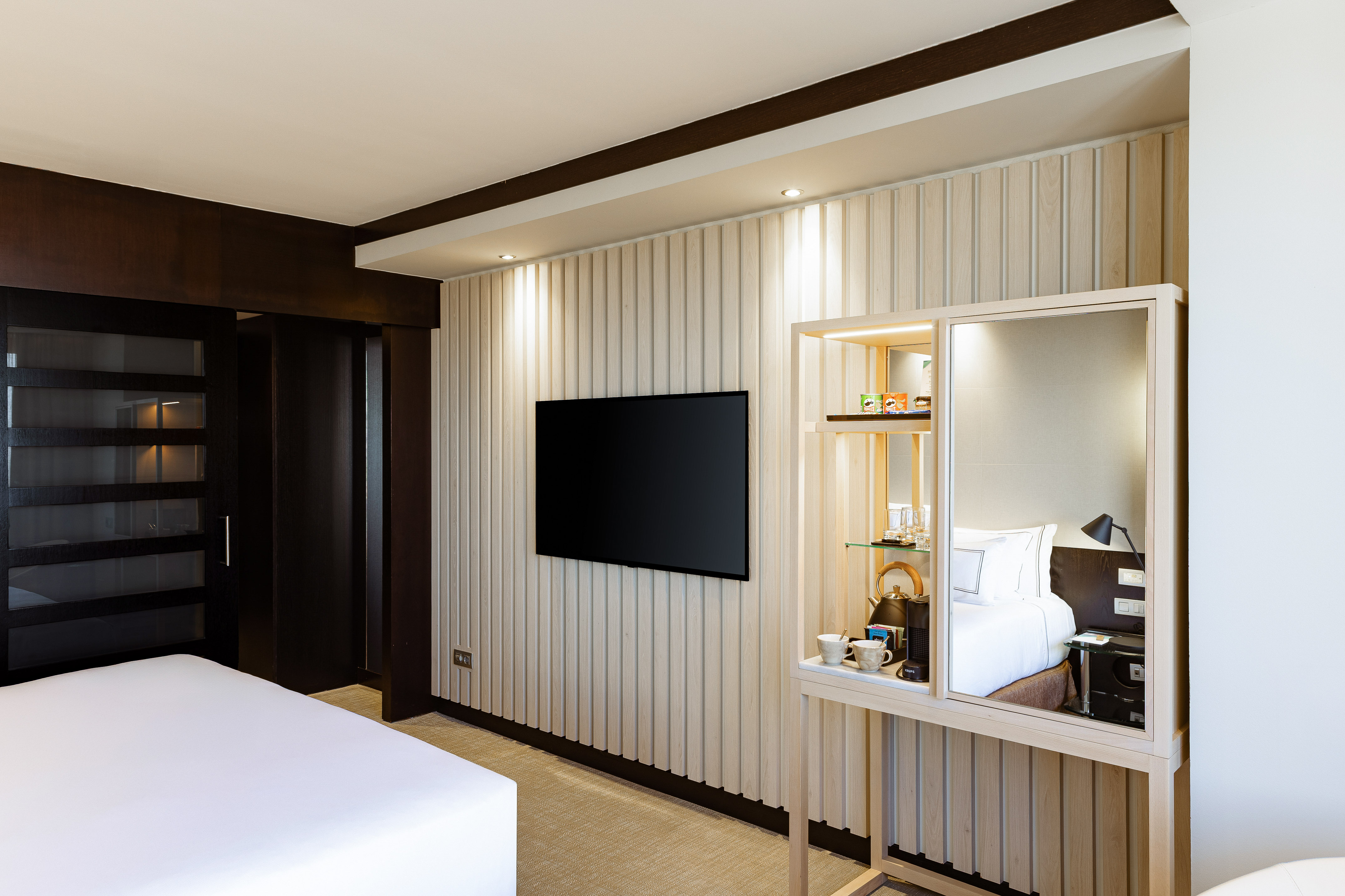 Contemporary hotel room with TV on ribbed wall, minibar, and white bed.