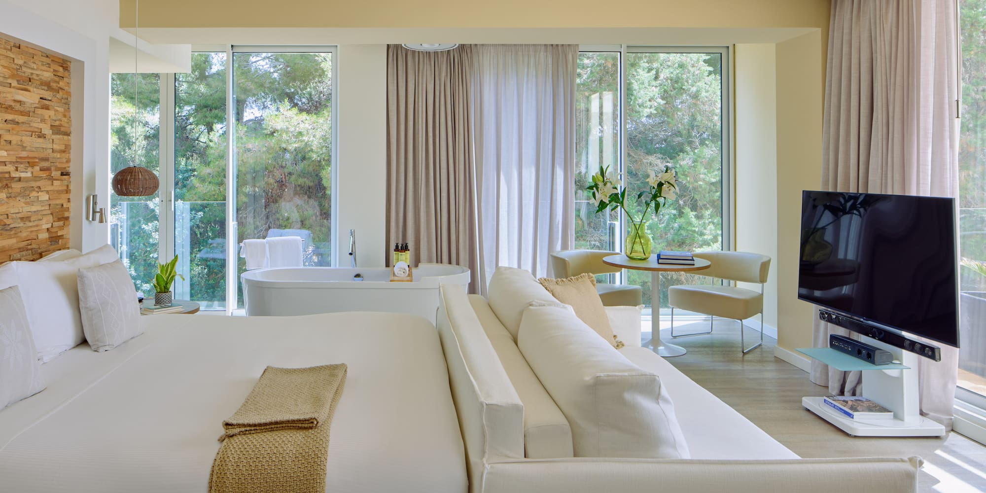 Luxury hotel suite with king bed, freestanding tub, sofa, and scenic nature view.