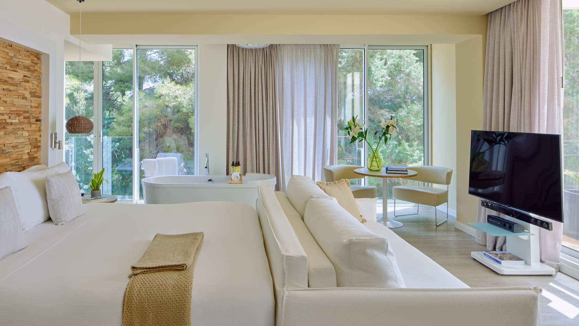 Luxury hotel suite with king bed, freestanding tub, sofa, and scenic nature view.