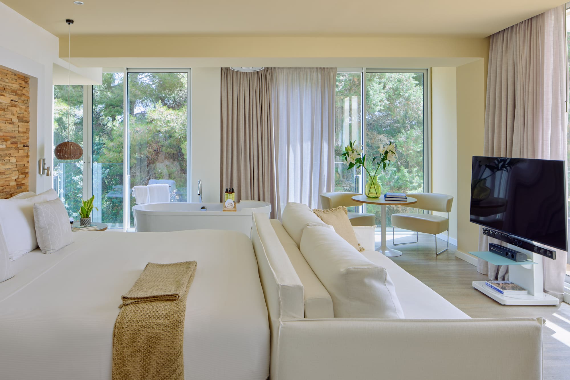 Luxury hotel suite with king bed, freestanding tub, sofa, and scenic nature view.