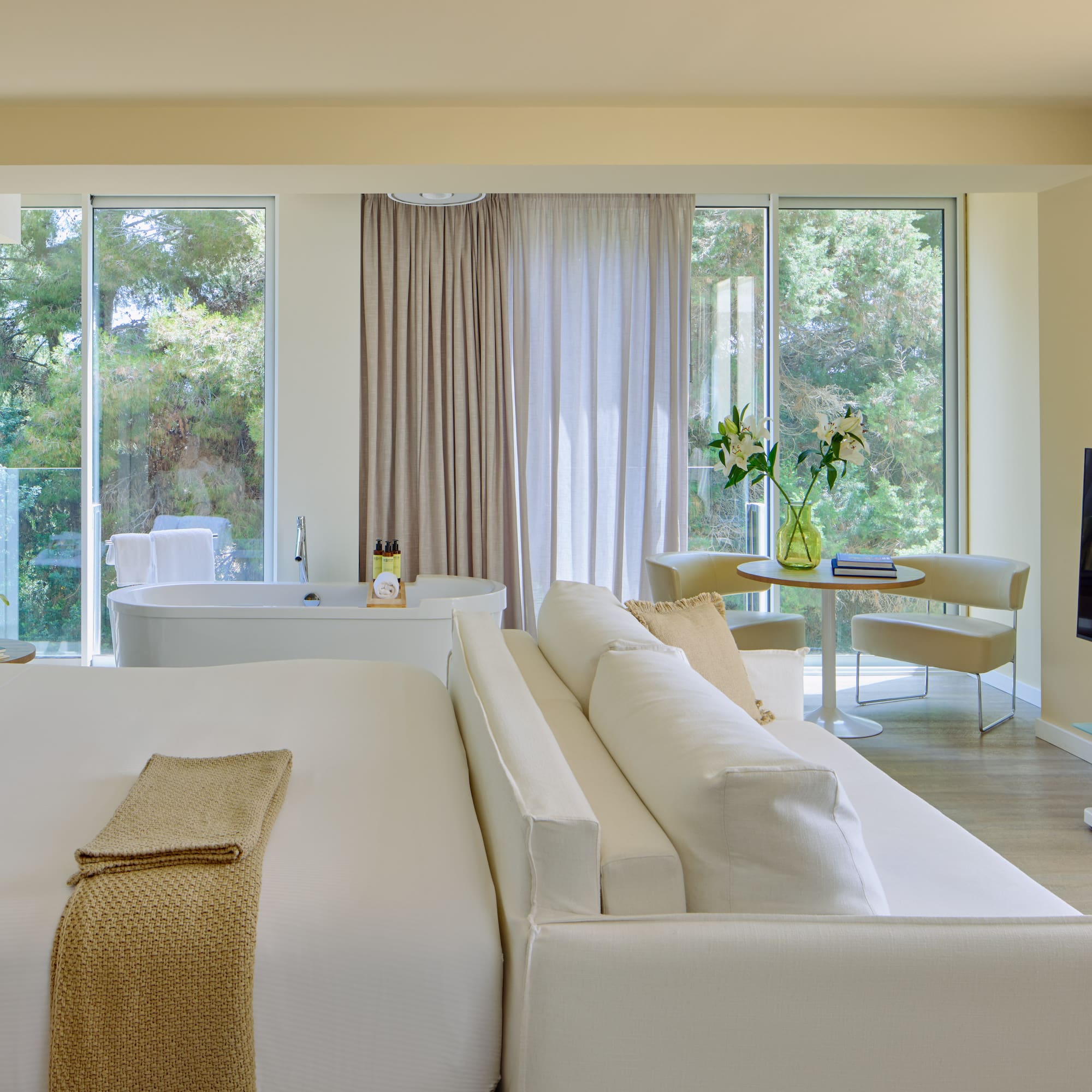 Luxury hotel suite with king bed, freestanding tub, sofa, and scenic nature view.