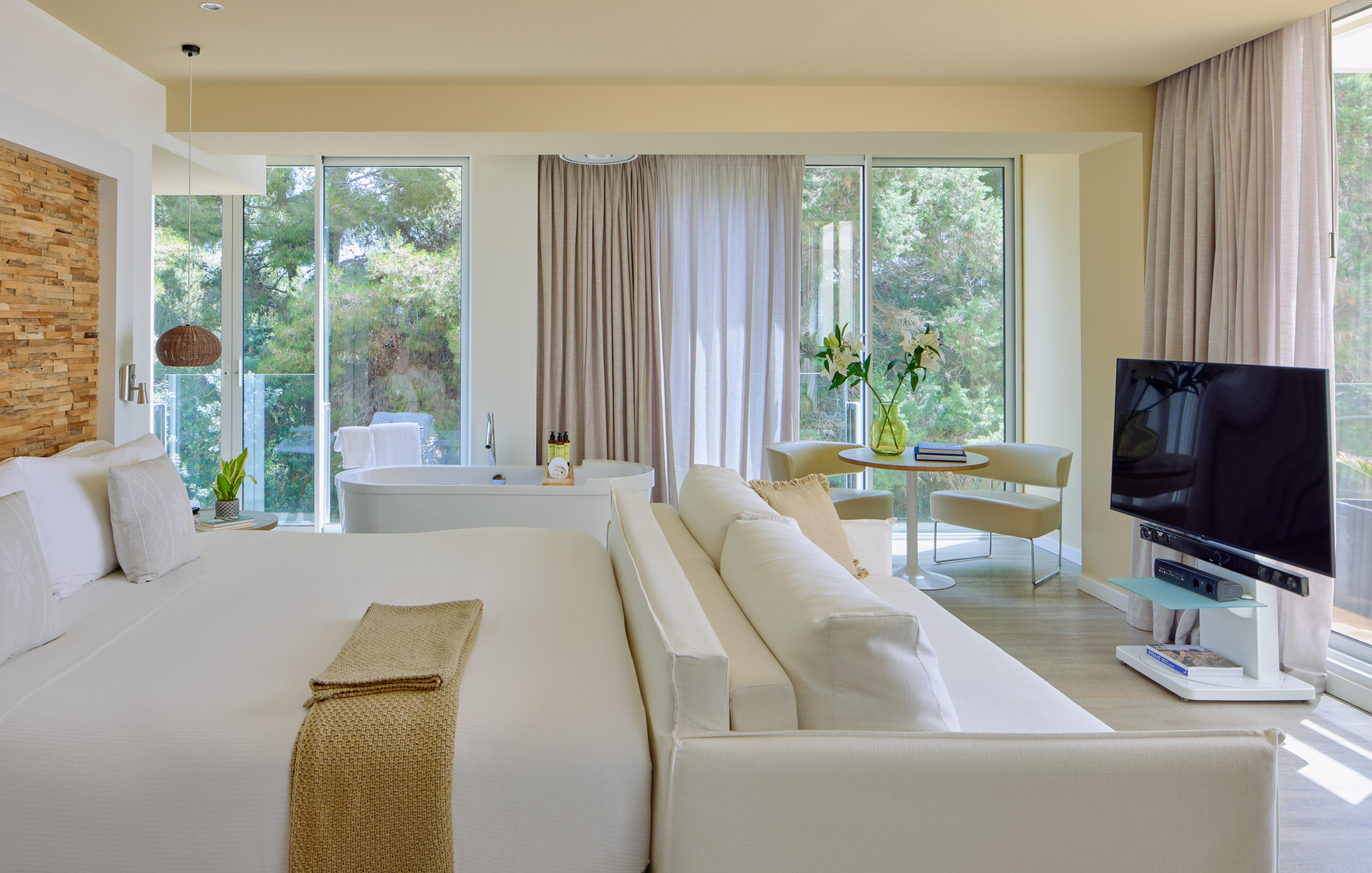 Luxury hotel suite with king bed, freestanding tub, sofa, and scenic nature view.