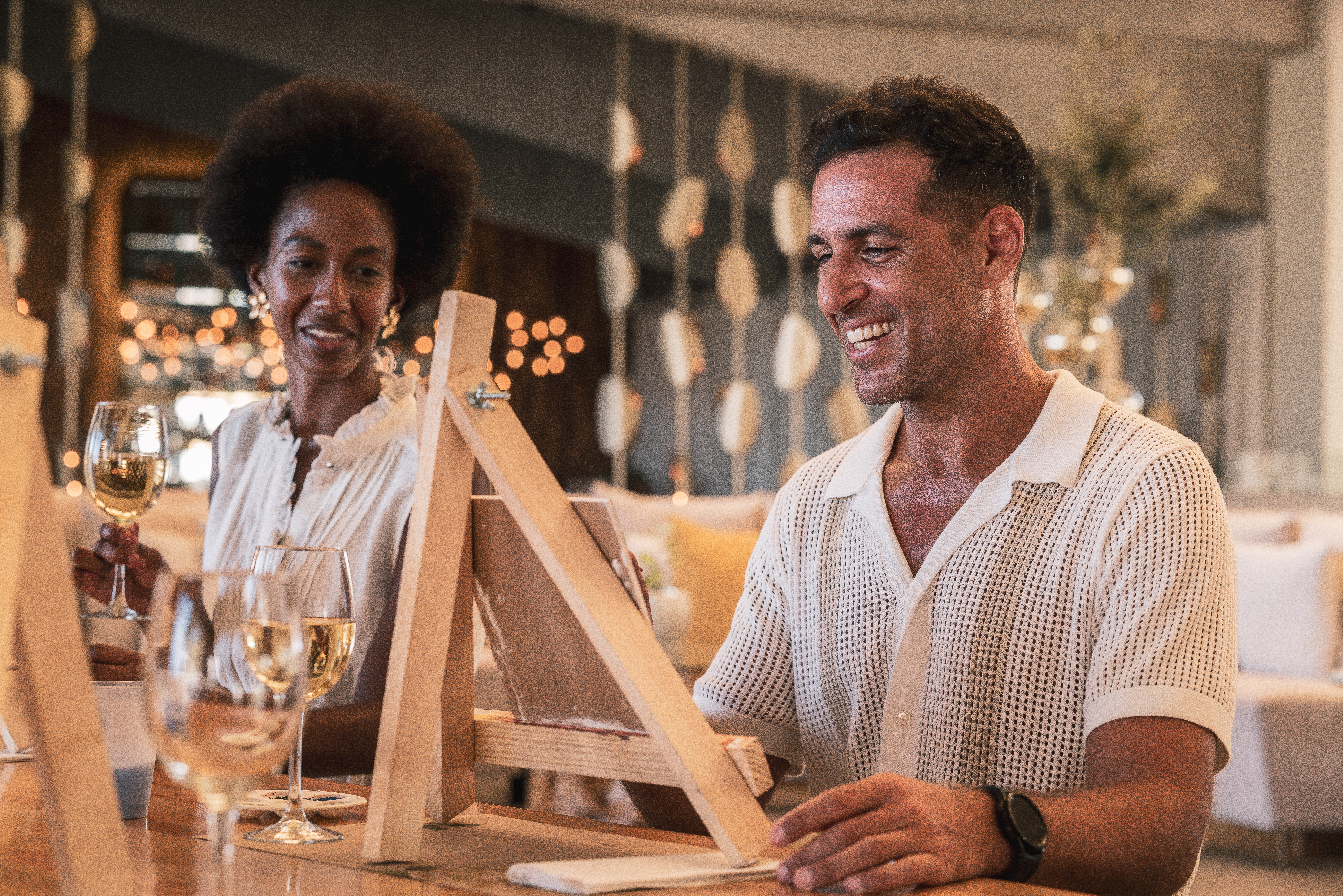 Smiling couple painting on easels, sipping wine at an art class.