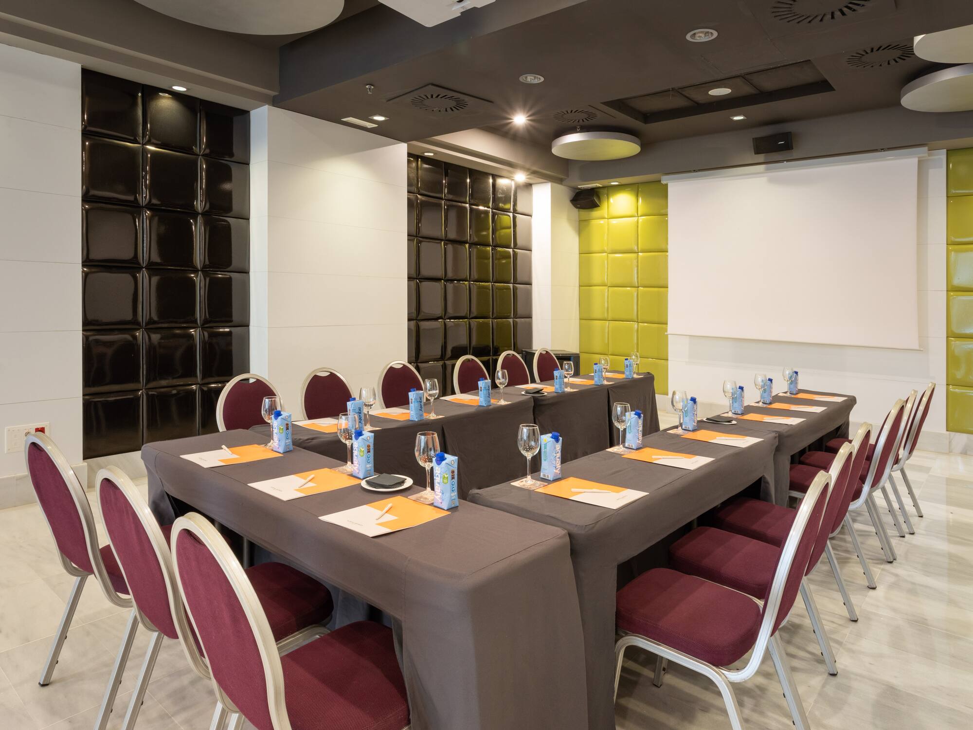 Modern conference room with U-shaped tables, maroon chairs, and projector screen.
