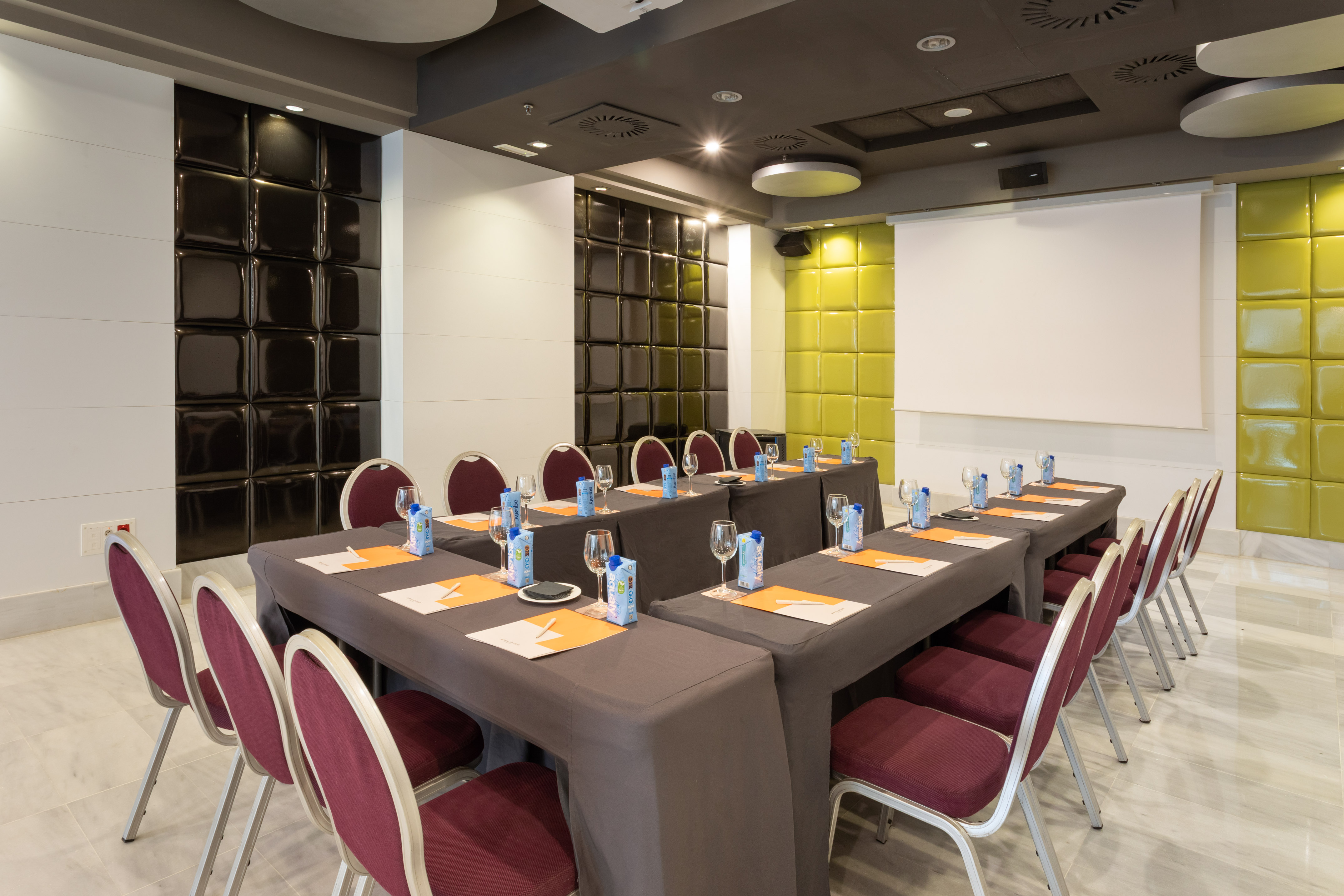 Modern conference room with U-shaped tables, maroon chairs, and projector screen.