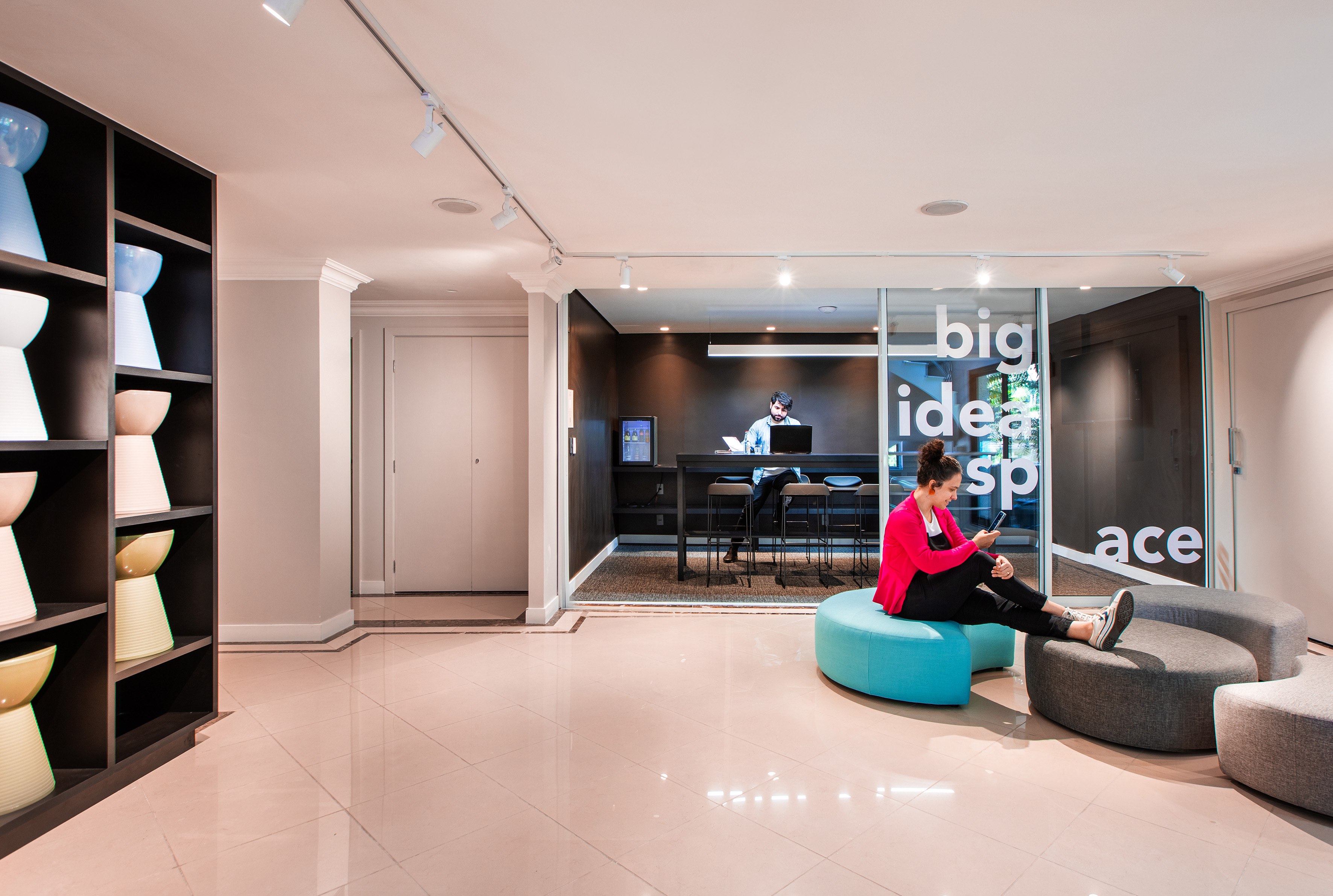 Modern office lounge with woman on phone, man working in glass-walled space.