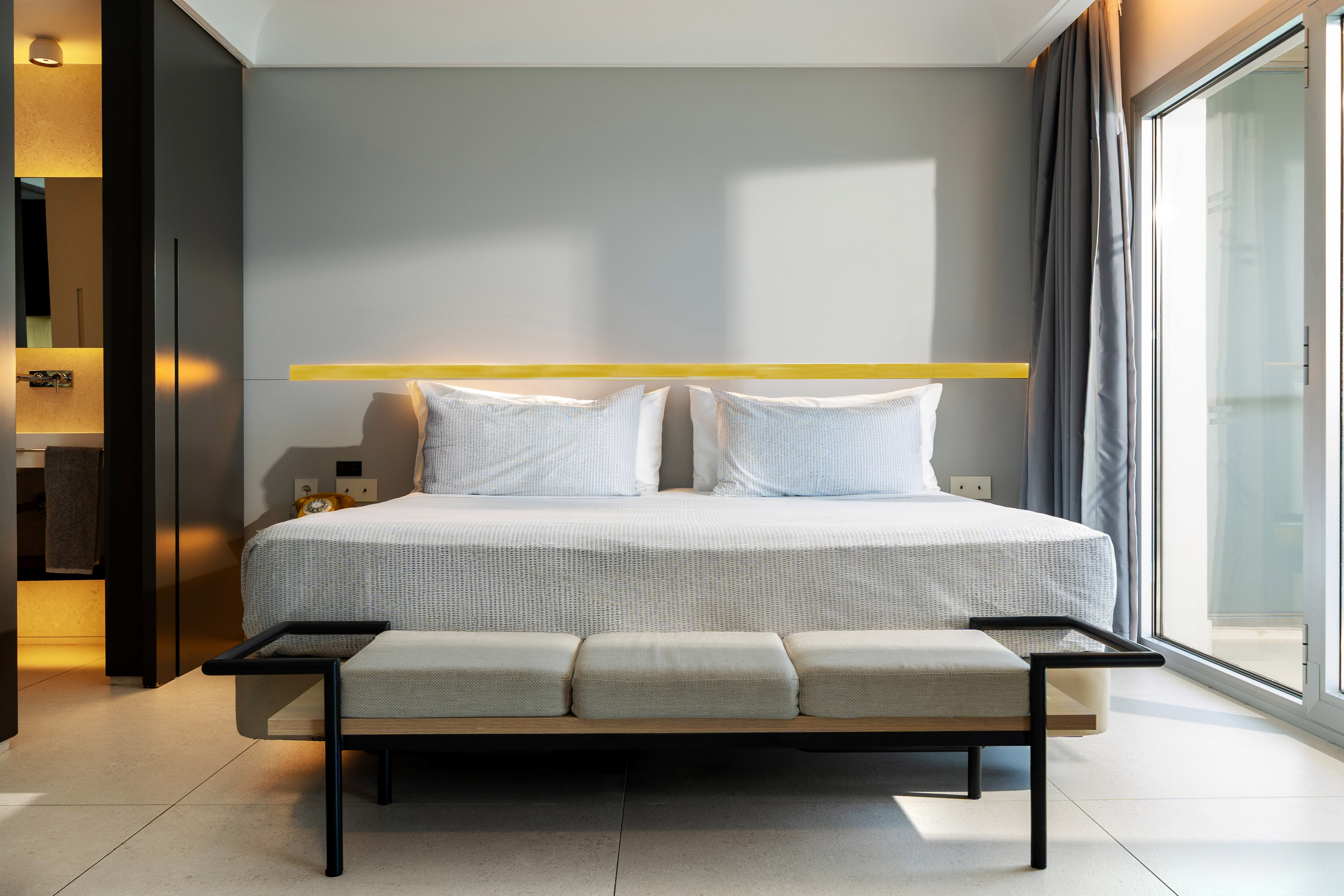 Modern hotel bedroom with king bed, warm yellow light, and glass door.