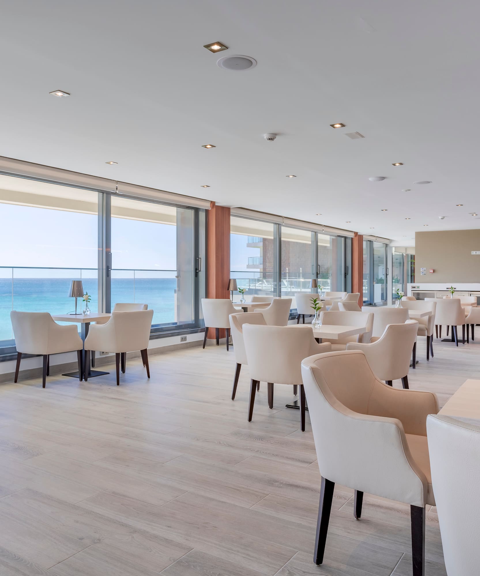 Restaurante moderno con vistas al mar, mesas y sillas claras y suelo de madera.