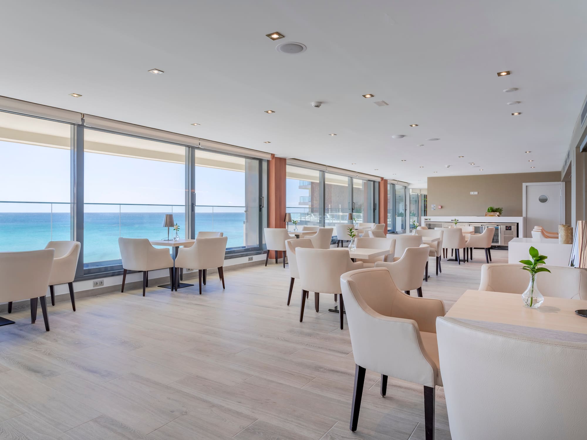 Restaurante moderno con vistas al mar, mesas y sillas claras y suelo de madera.