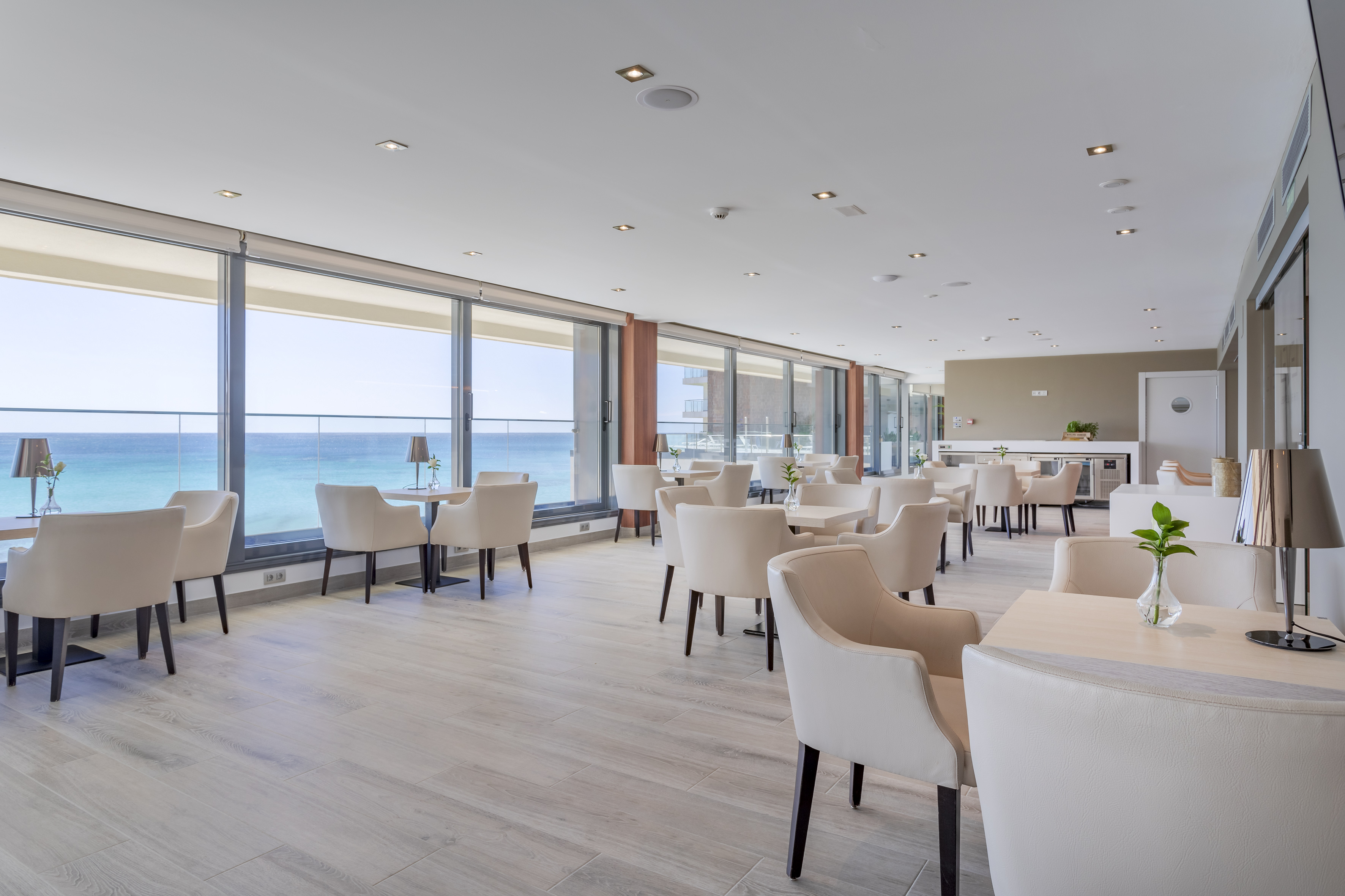 Restaurante moderno con vistas al mar, mesas y sillas claras y suelo de madera.