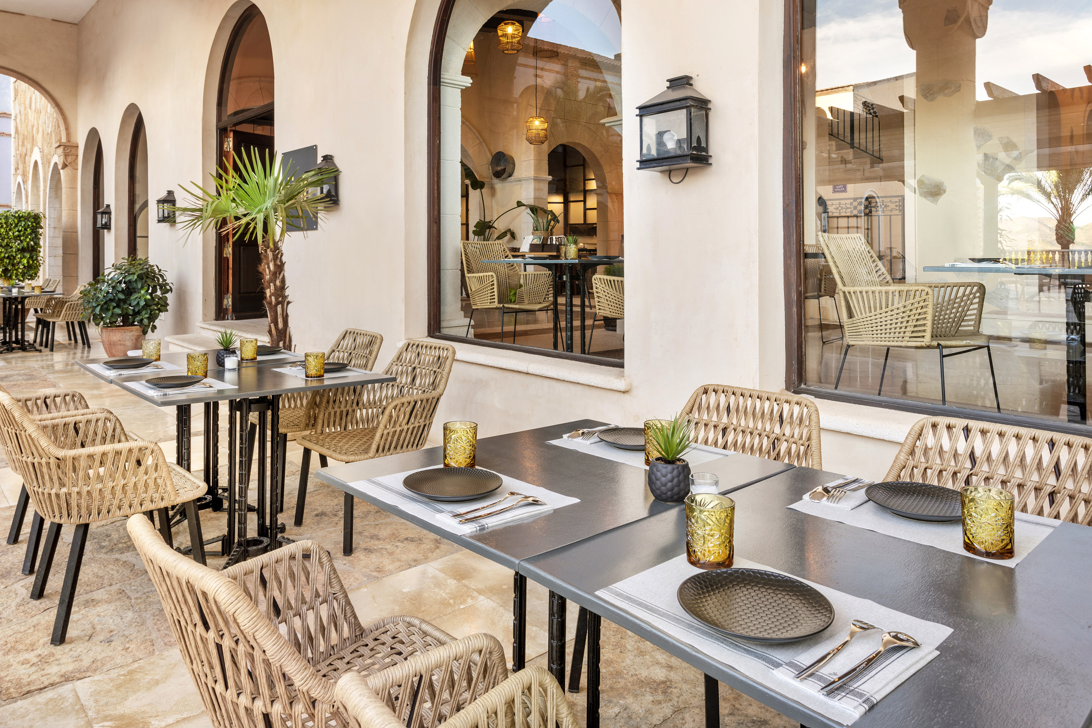 Outdoor restaurant patio with tables, wicker chairs, and arched architecture.
