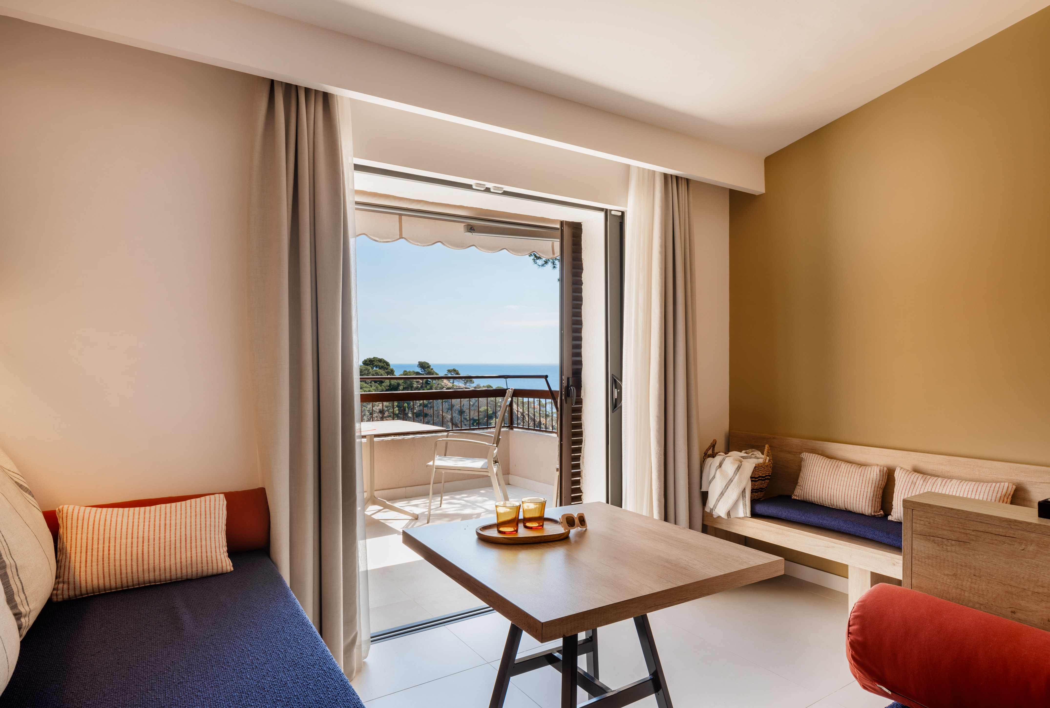 Modern hotel room with sea view balcony, wooden table, and comfortable seating.