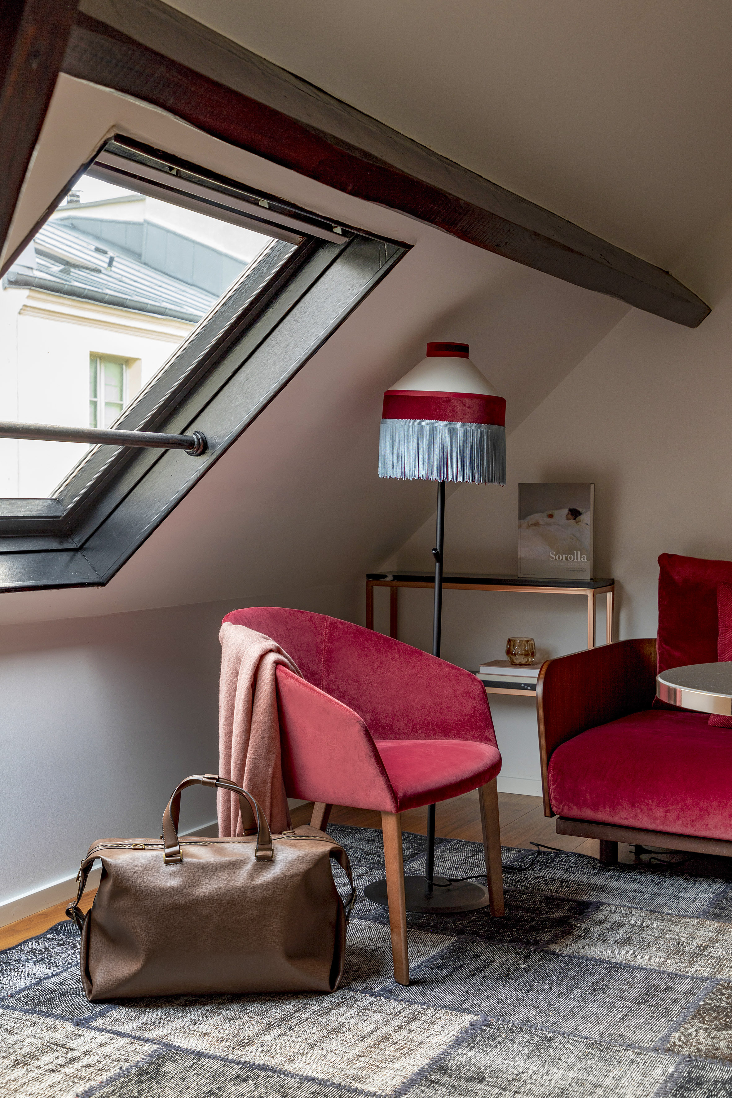 Cozy attic room with skylight, red velvet chairs, fringed lamp, and travel bag.