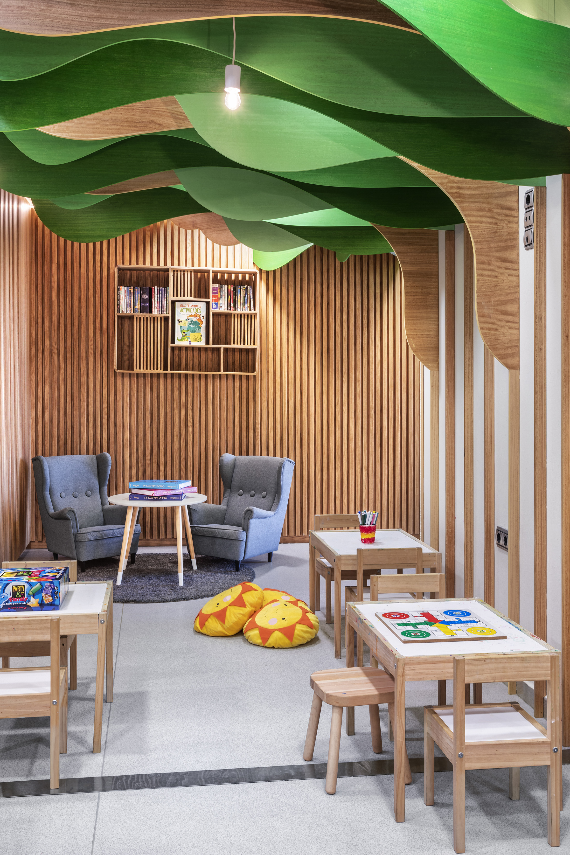 Kids' reading nook with green leaf ceiling, wooden walls, small tables, chairs, books.