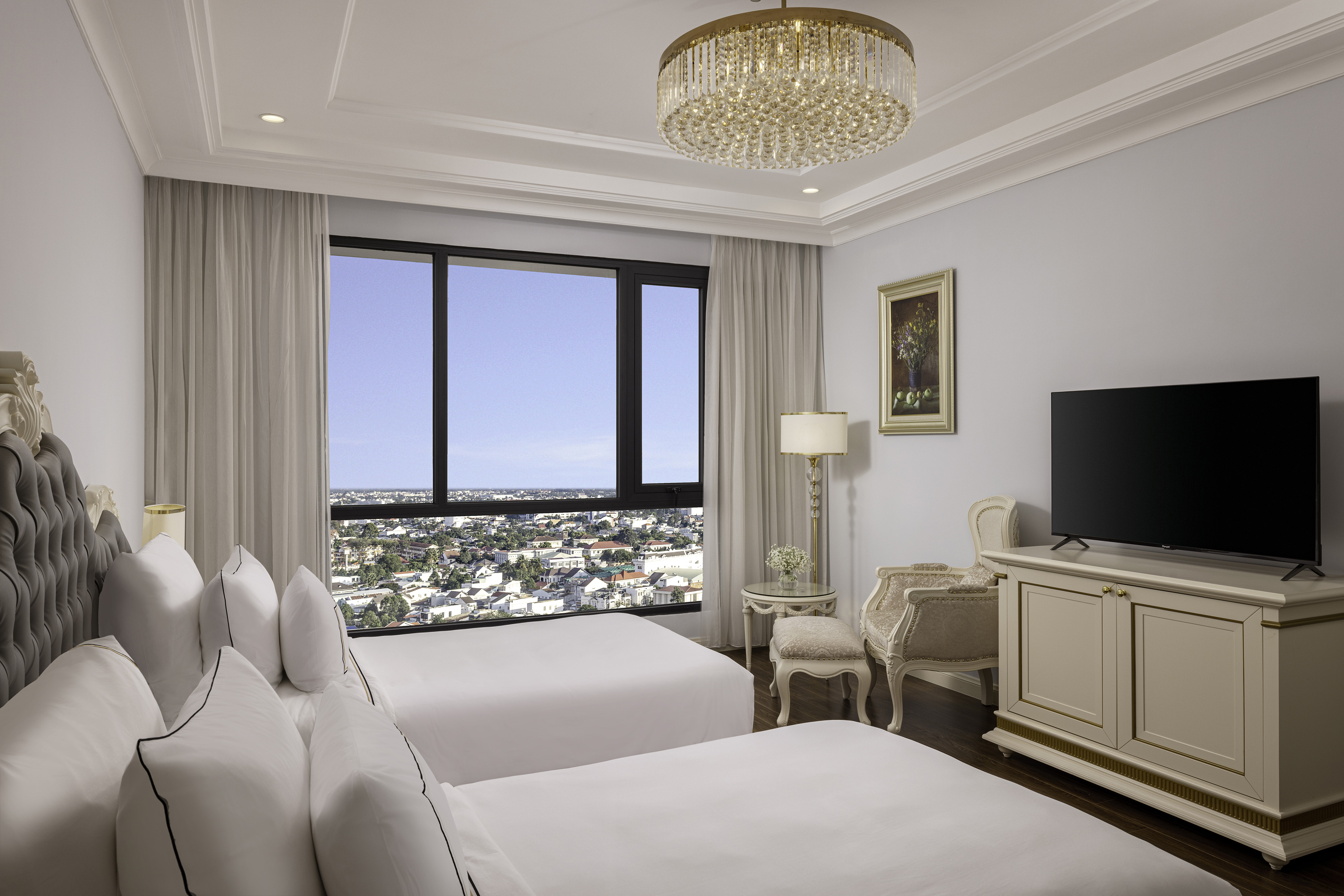 Luxury hotel room with twin beds, city view, chandelier, and elegant decor.