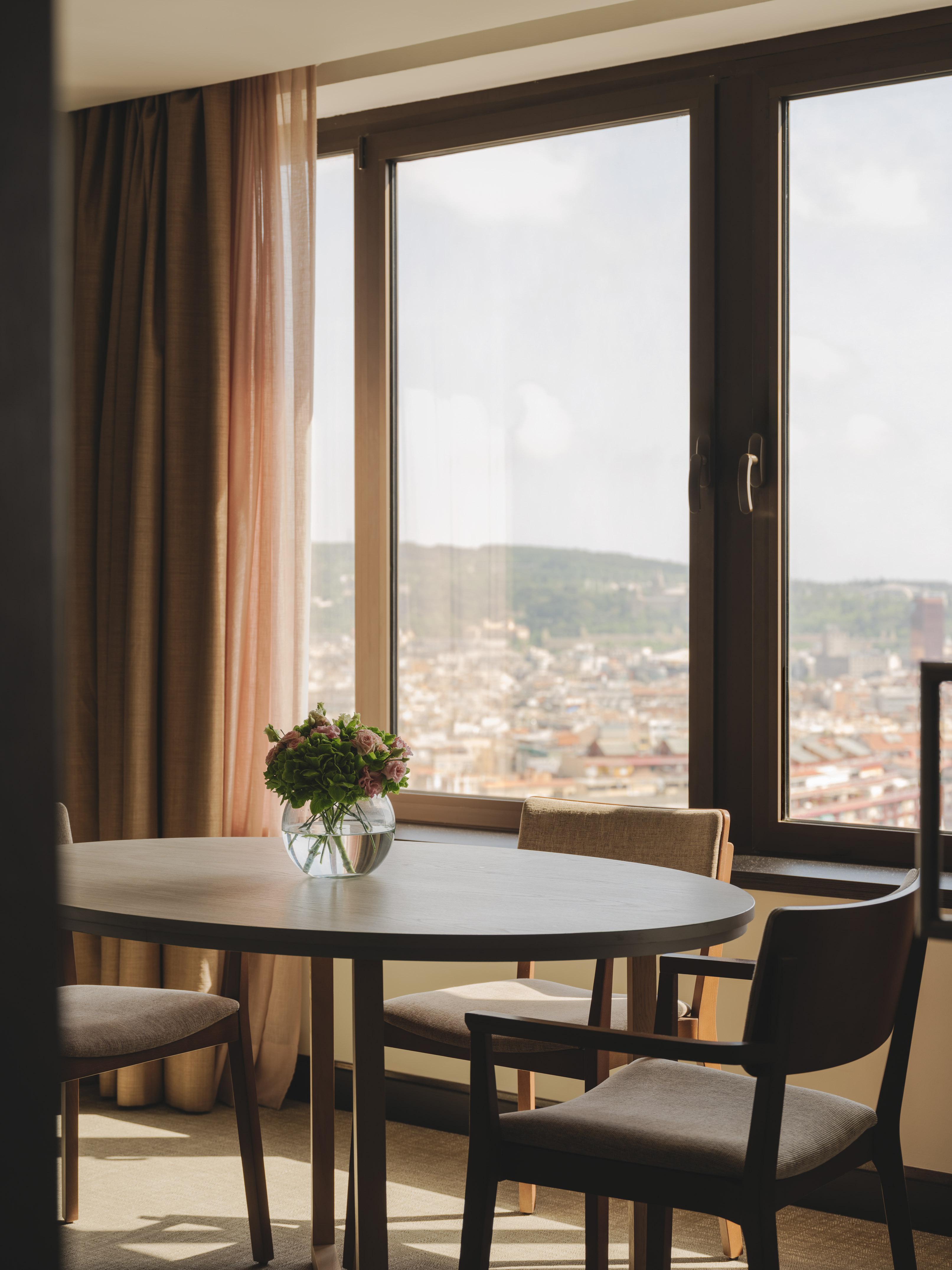 Mesa redonda, jarrón con flores, ventanal con vistas a la ciudad.