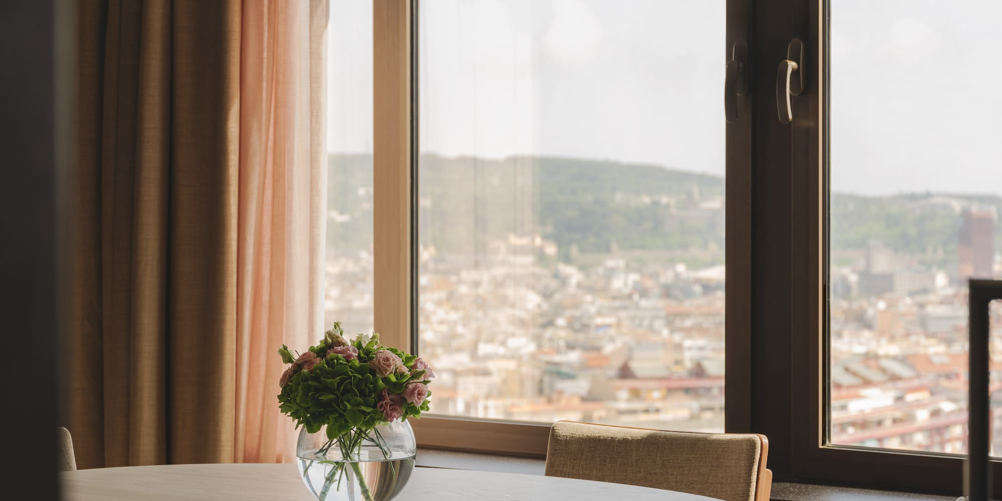 Mesa redonda, jarrón con flores, ventanal con vistas a la ciudad.