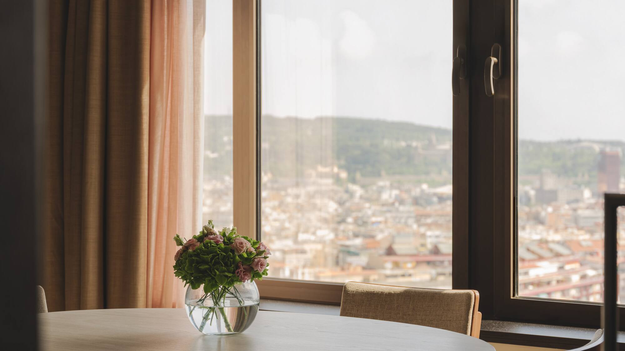 Mesa redonda, jarrón con flores, ventanal con vistas a la ciudad.