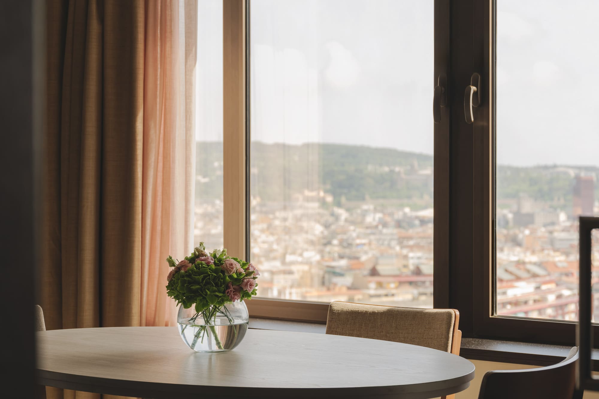 Mesa redonda, jarrón con flores, ventanal con vistas a la ciudad.