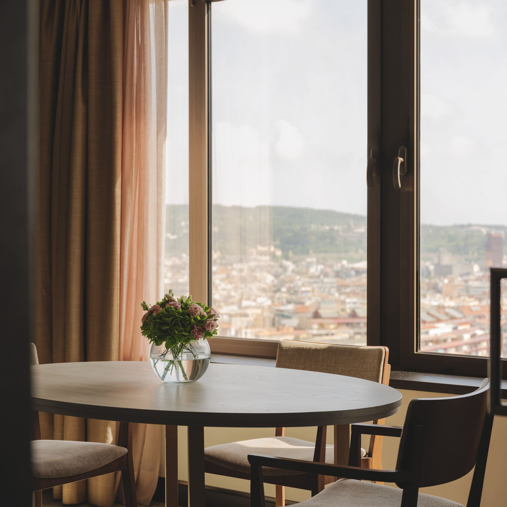 Mesa redonda, jarrón con flores, ventanal con vistas a la ciudad.