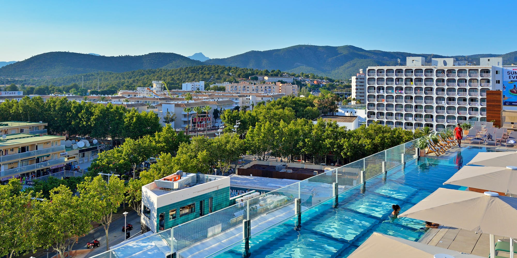 Piscina transparente en azotea de hotel con vistas a ciudad y montañas soleadas.
