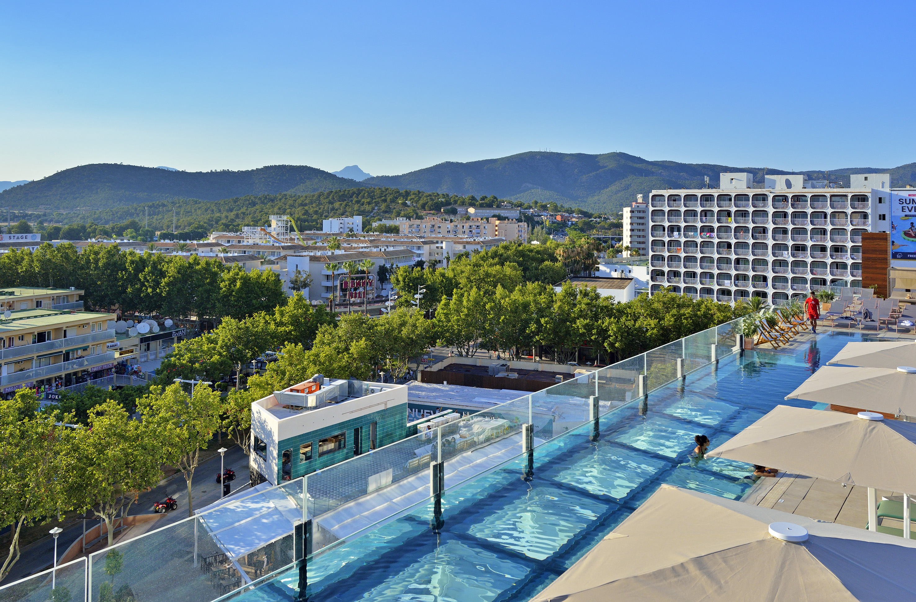 Piscina transparente en azotea de hotel con vistas a ciudad y montañas soleadas.