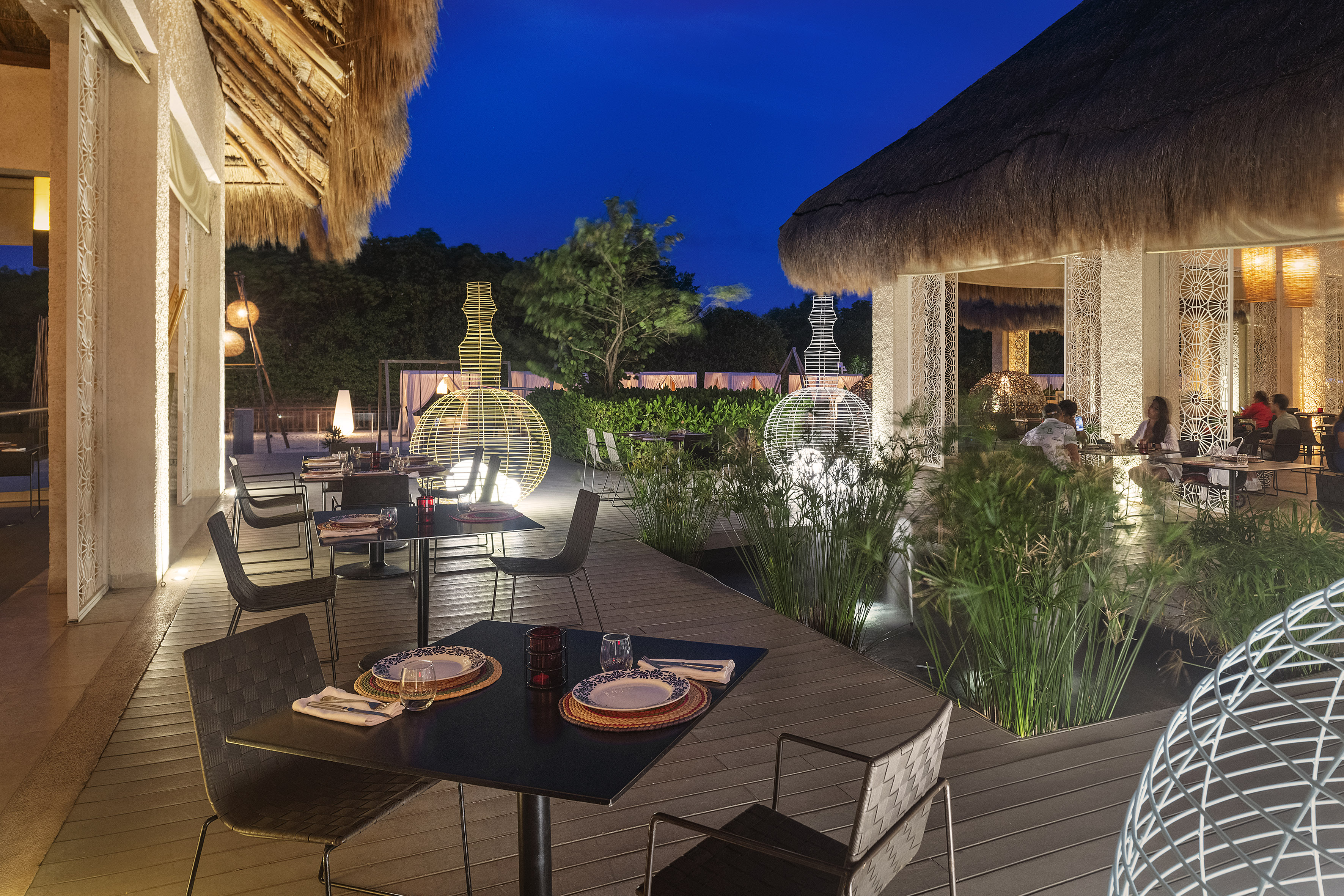 Thatched roof outdoor dining, night, glowing lights, elegant tables.