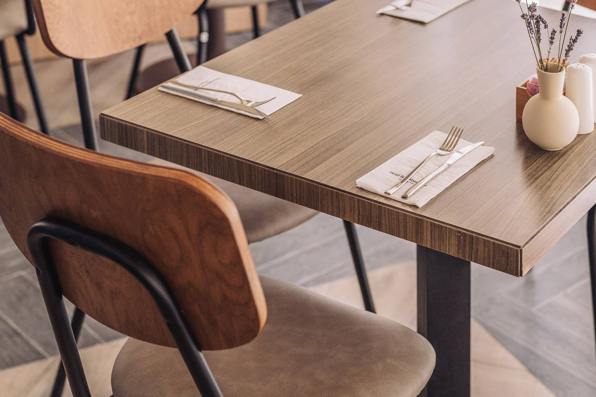 Modern restaurant dining table set with cutlery, lavender vase, and wooden chairs.