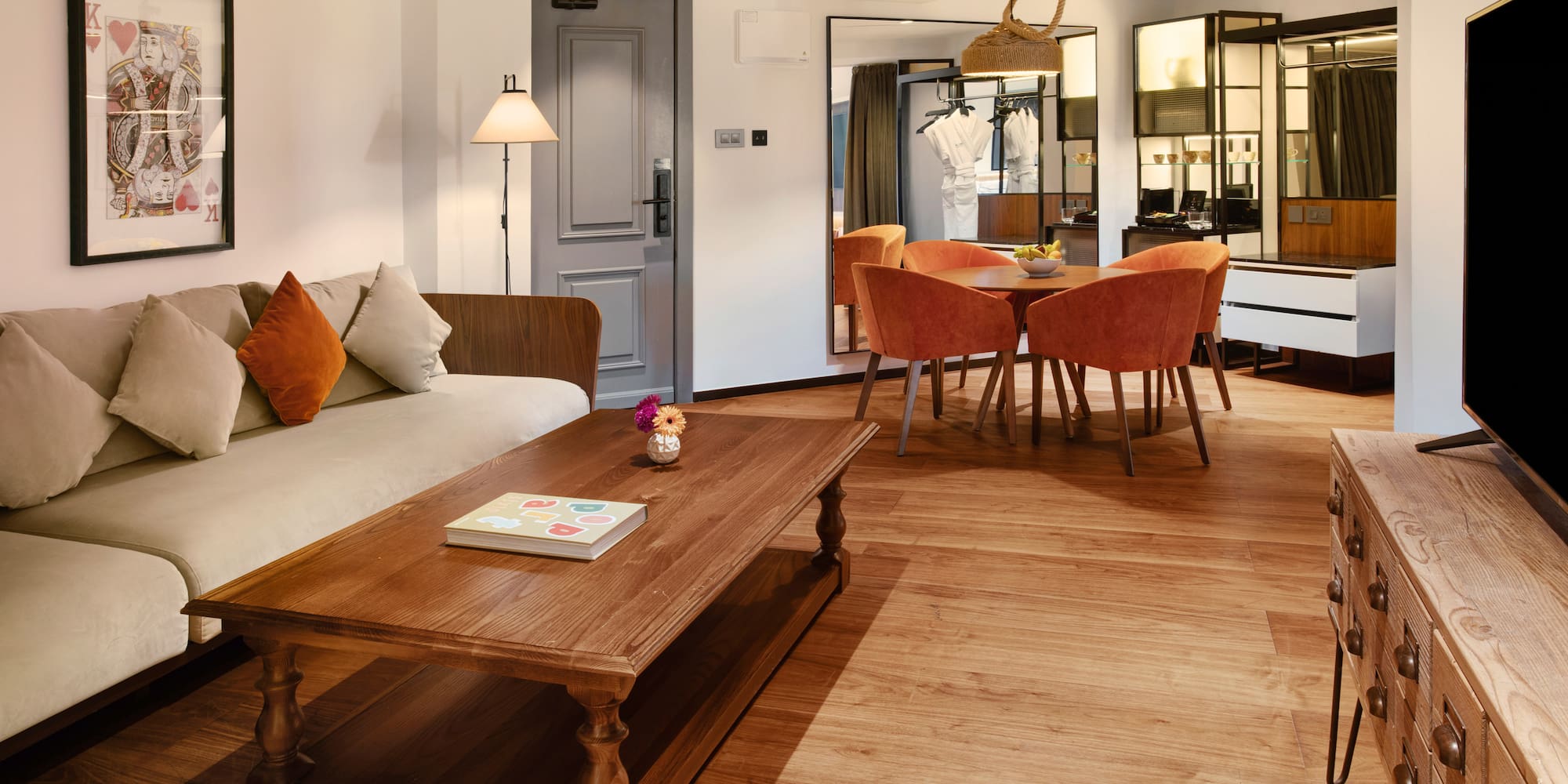 Modern hotel suite living-dining area with sofa, wooden table, orange chairs.