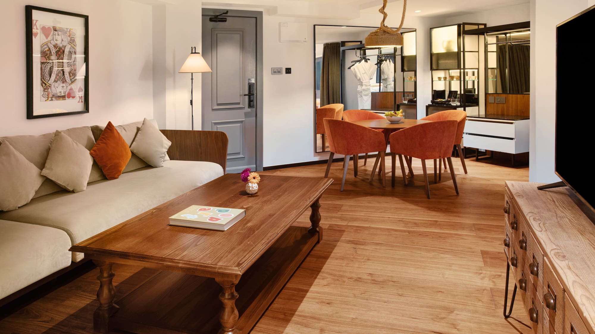 Modern hotel suite living-dining area with sofa, wooden table, orange chairs.
