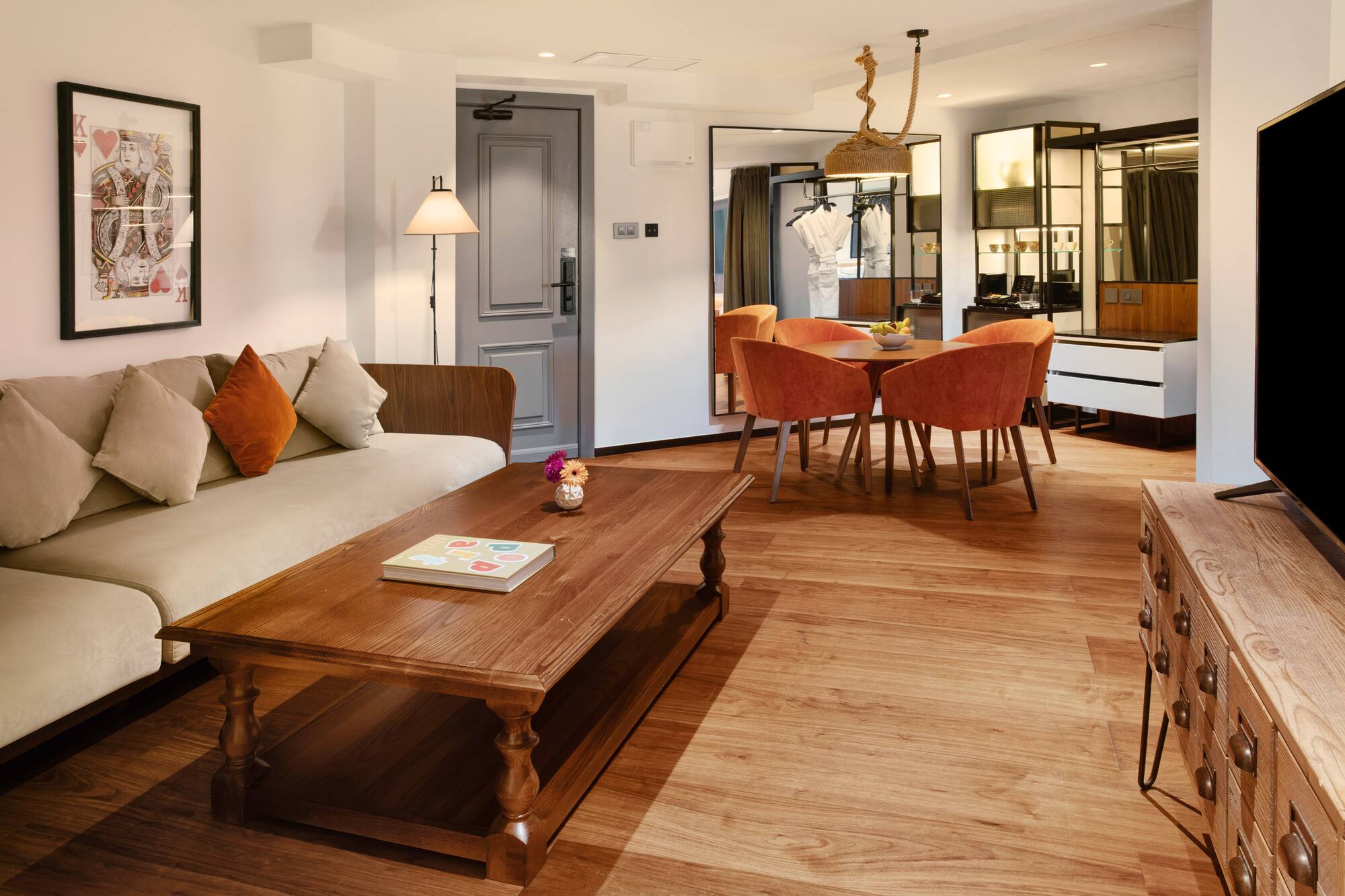 Modern hotel suite living-dining area with sofa, wooden table, orange chairs.