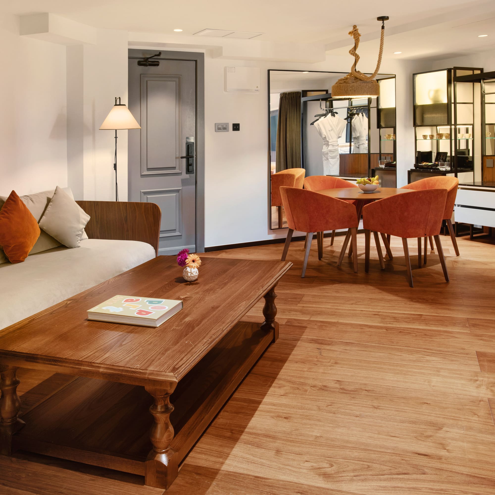 Modern hotel suite living-dining area with sofa, wooden table, orange chairs.