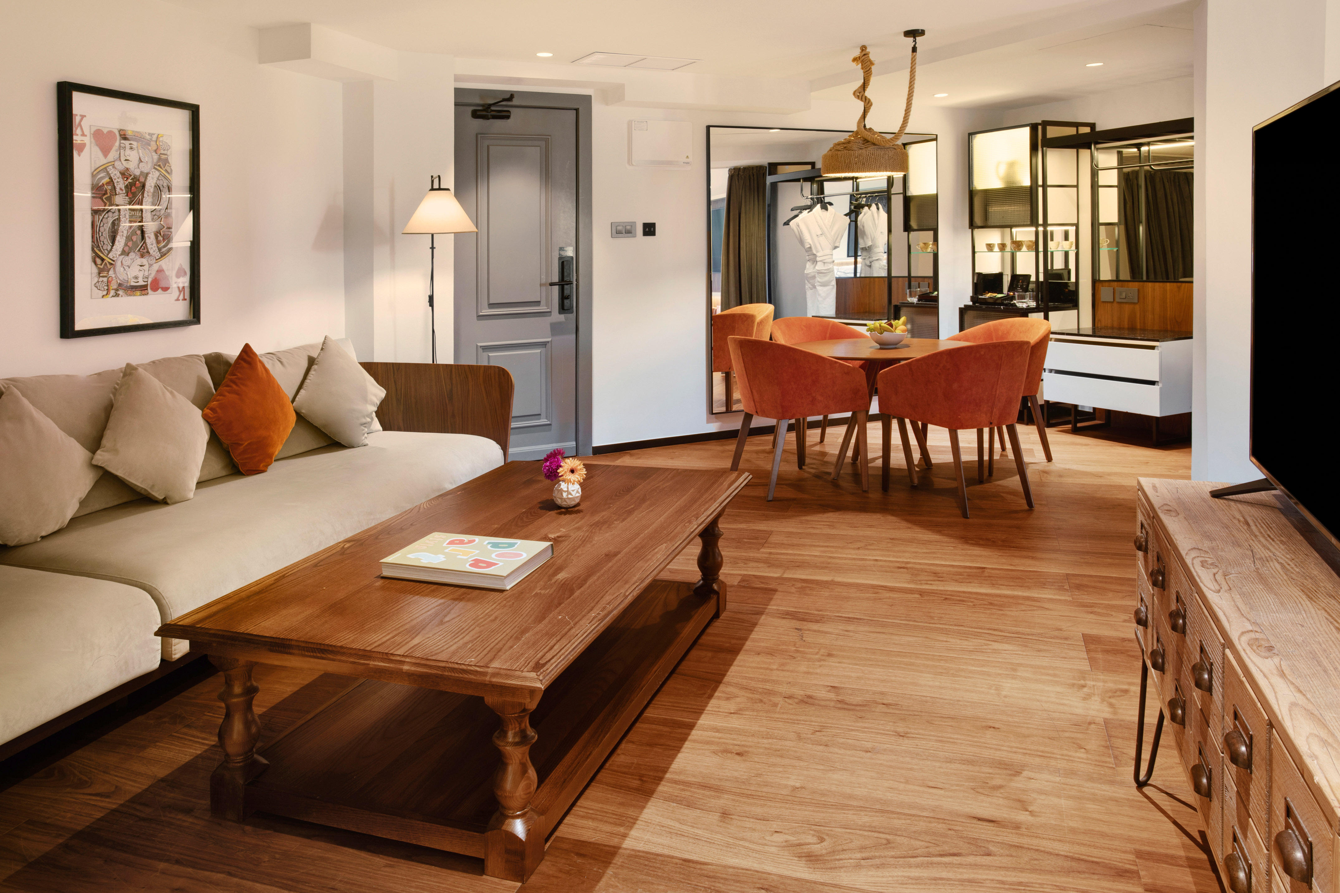 Modern hotel suite living-dining area with sofa, wooden table, orange chairs.