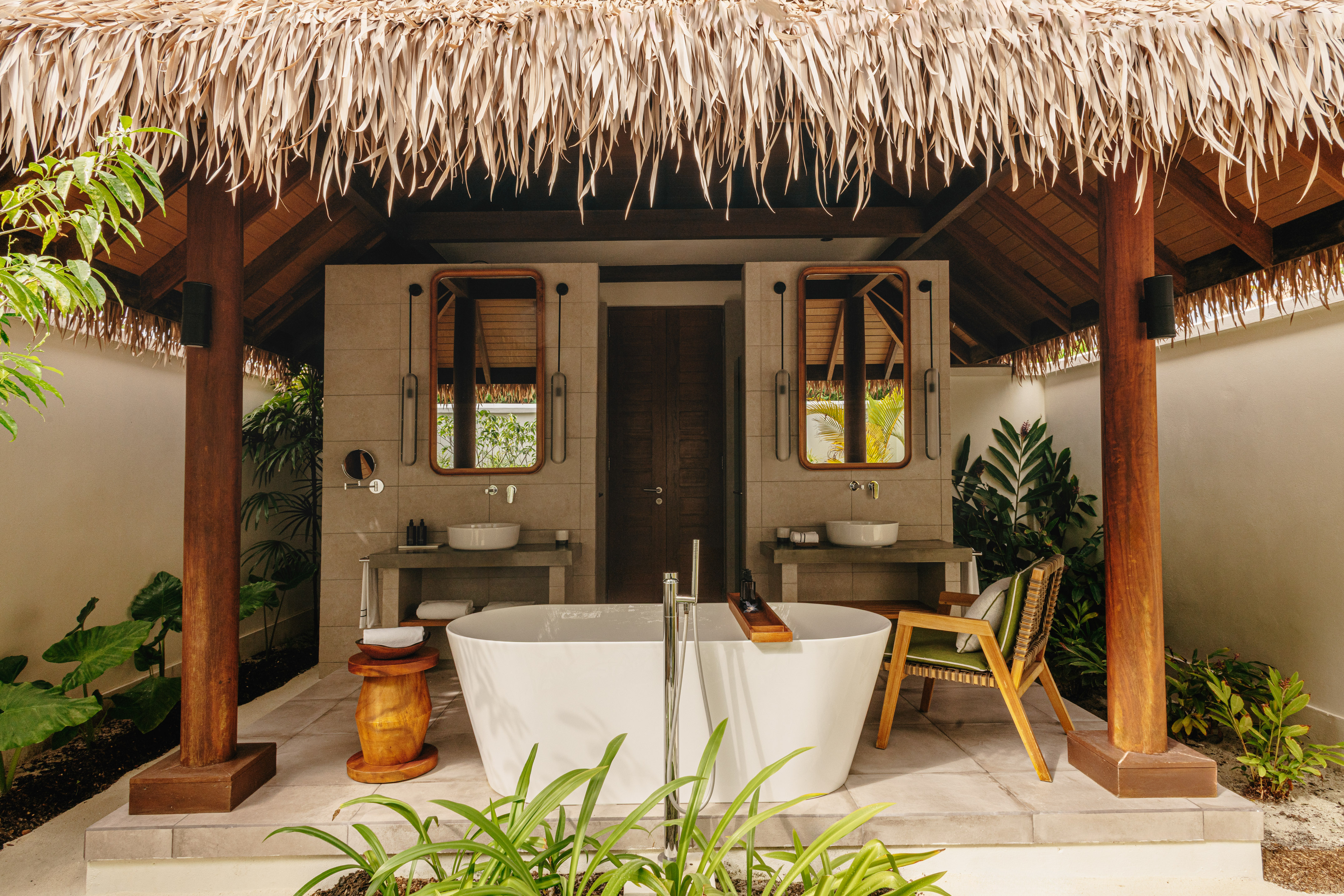 Outdoor bathroom with freestanding tub, twin sinks, thatched roof, and tropical plants.