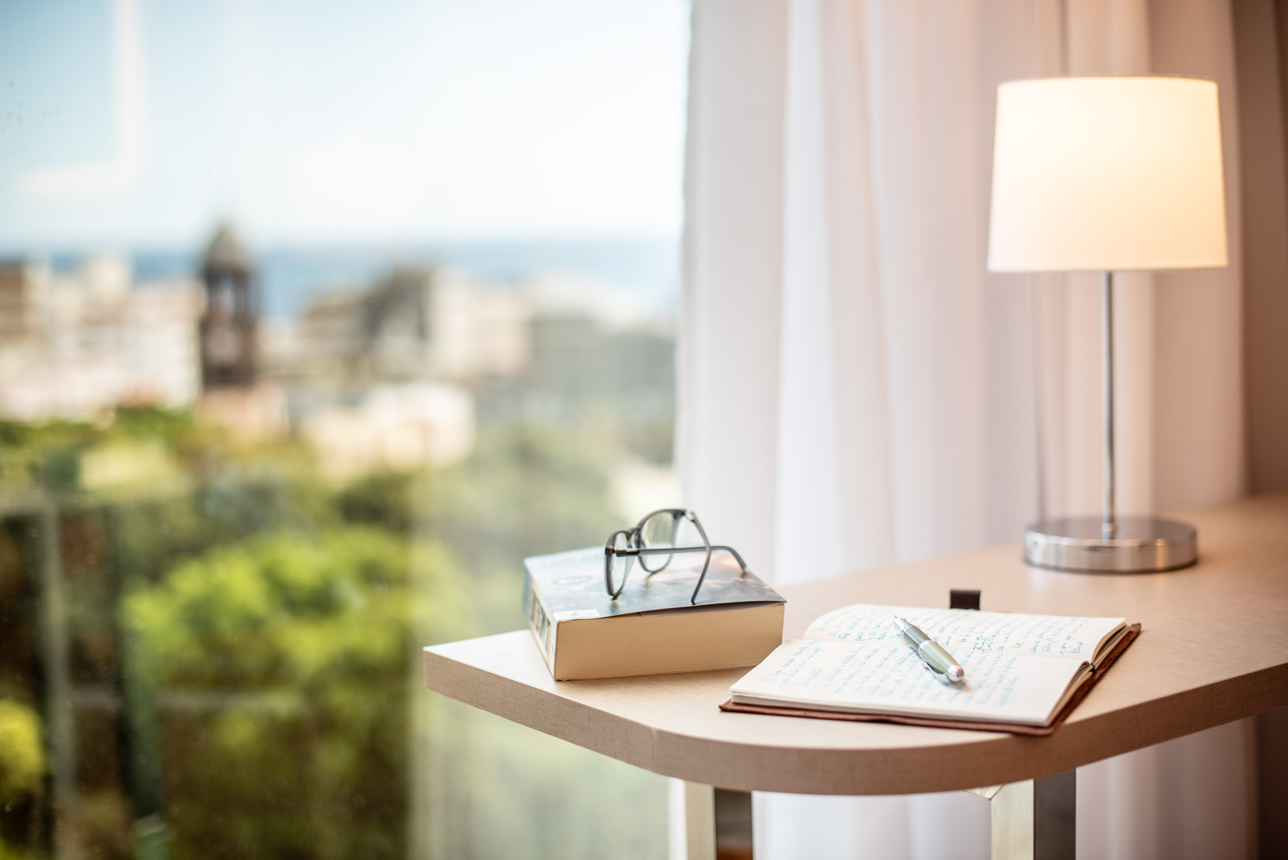 Study desk with book, glasses, notebook, lamp by window, city-ocean view.