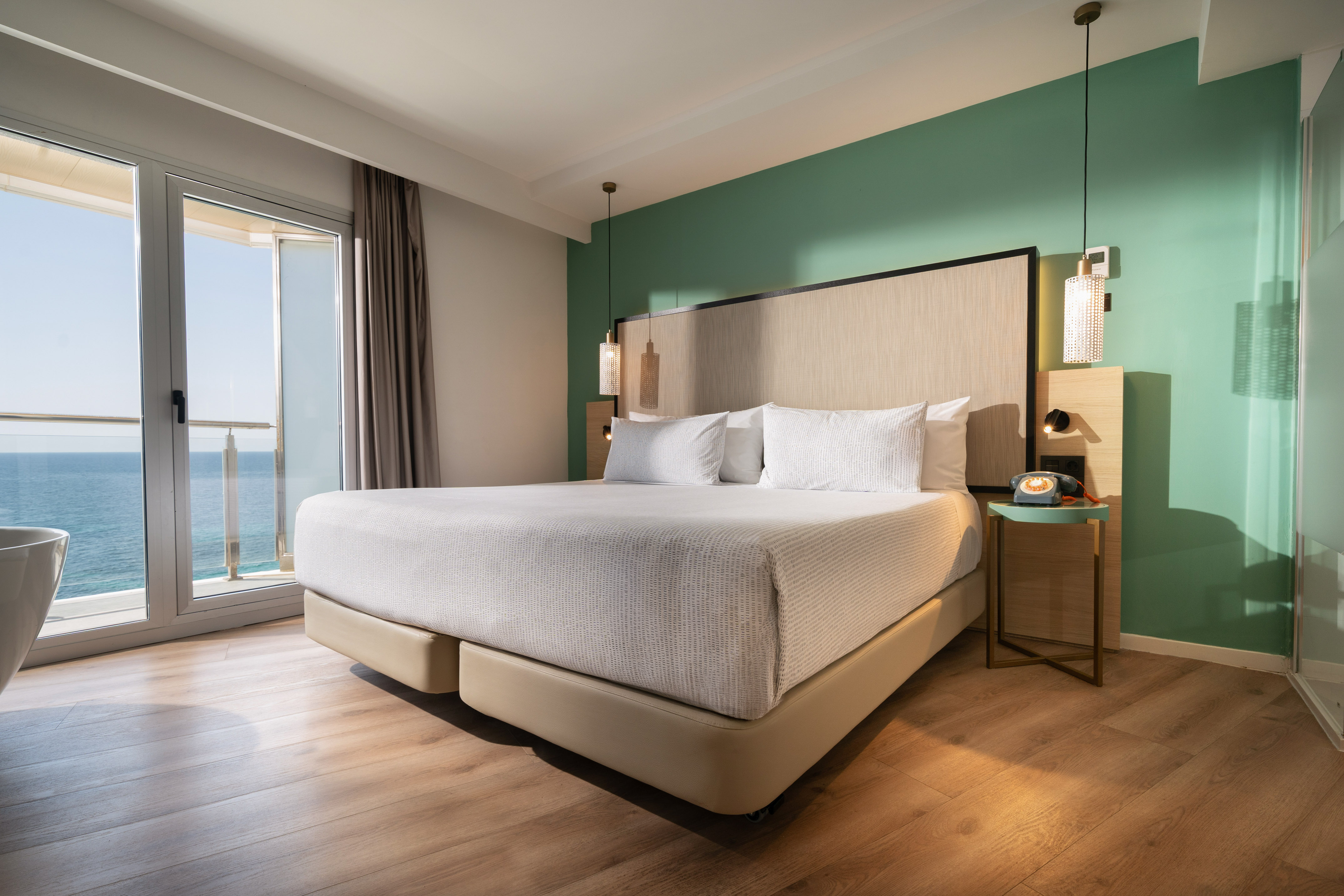 Modern hotel room with king bed, sea view balcony, and green accent wall.