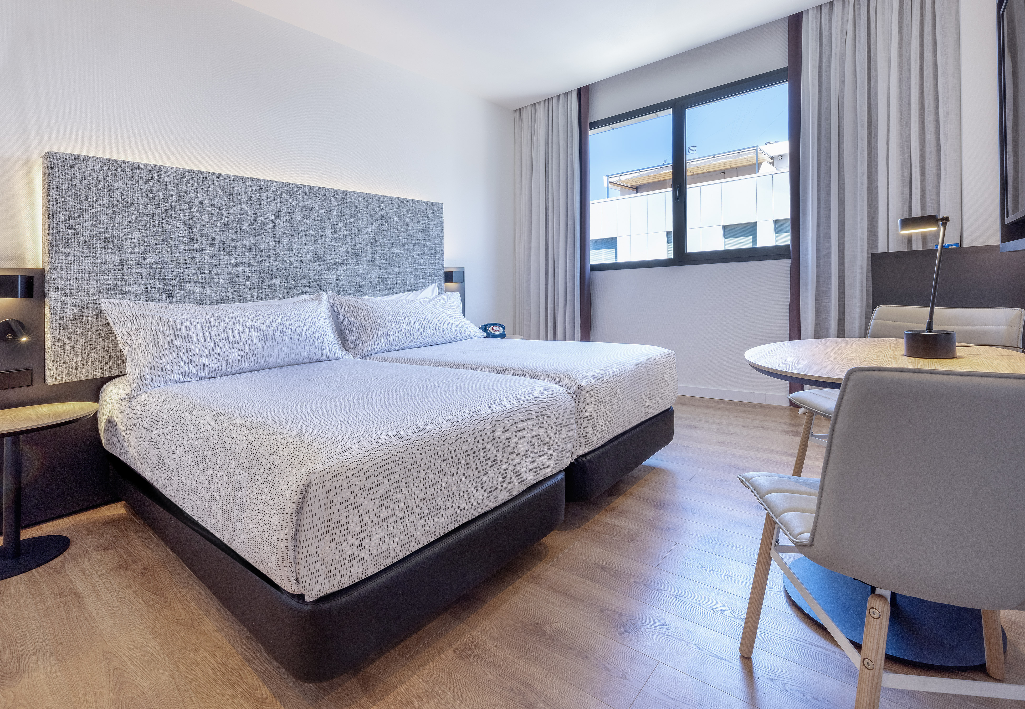 Bright modern hotel room with twin beds, wood floor, window, and dining table.