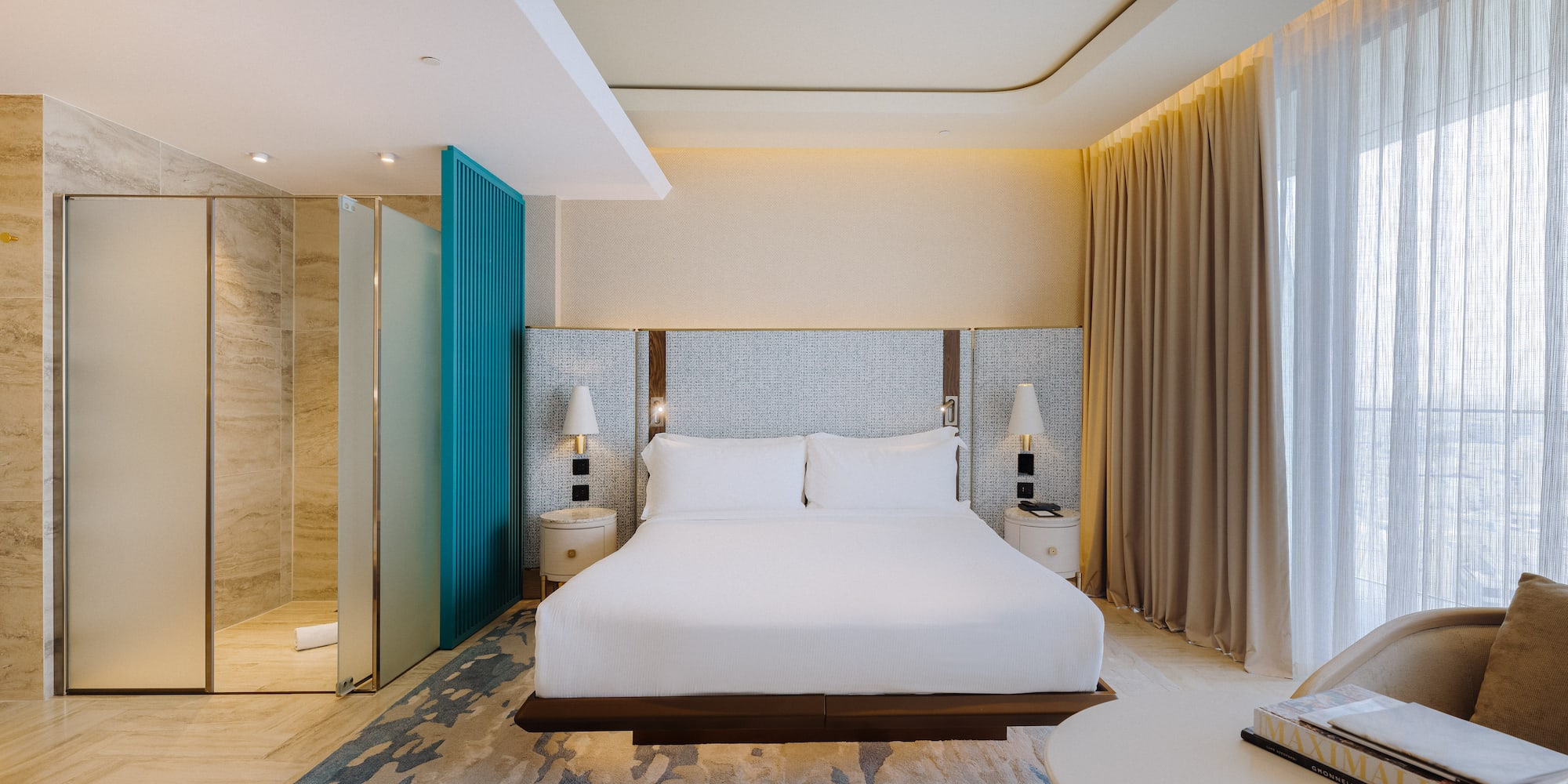 Modern hotel room with king bed, glass shower, teal accent, and sheer curtains.