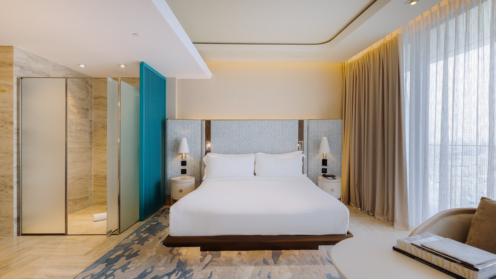 Modern hotel room with king bed, glass shower, teal accent, and sheer curtains.