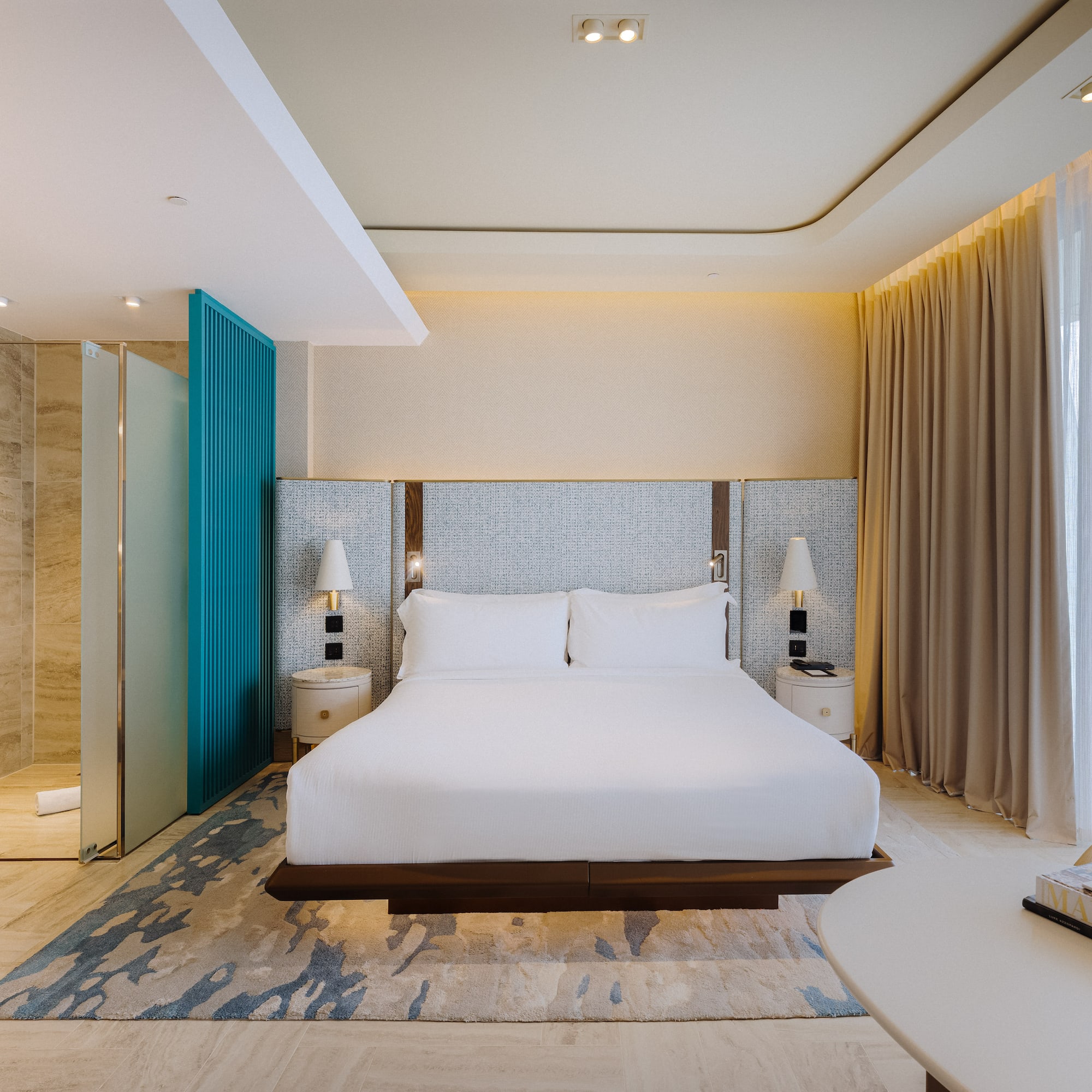 Modern hotel room with king bed, glass shower, teal accent, and sheer curtains.