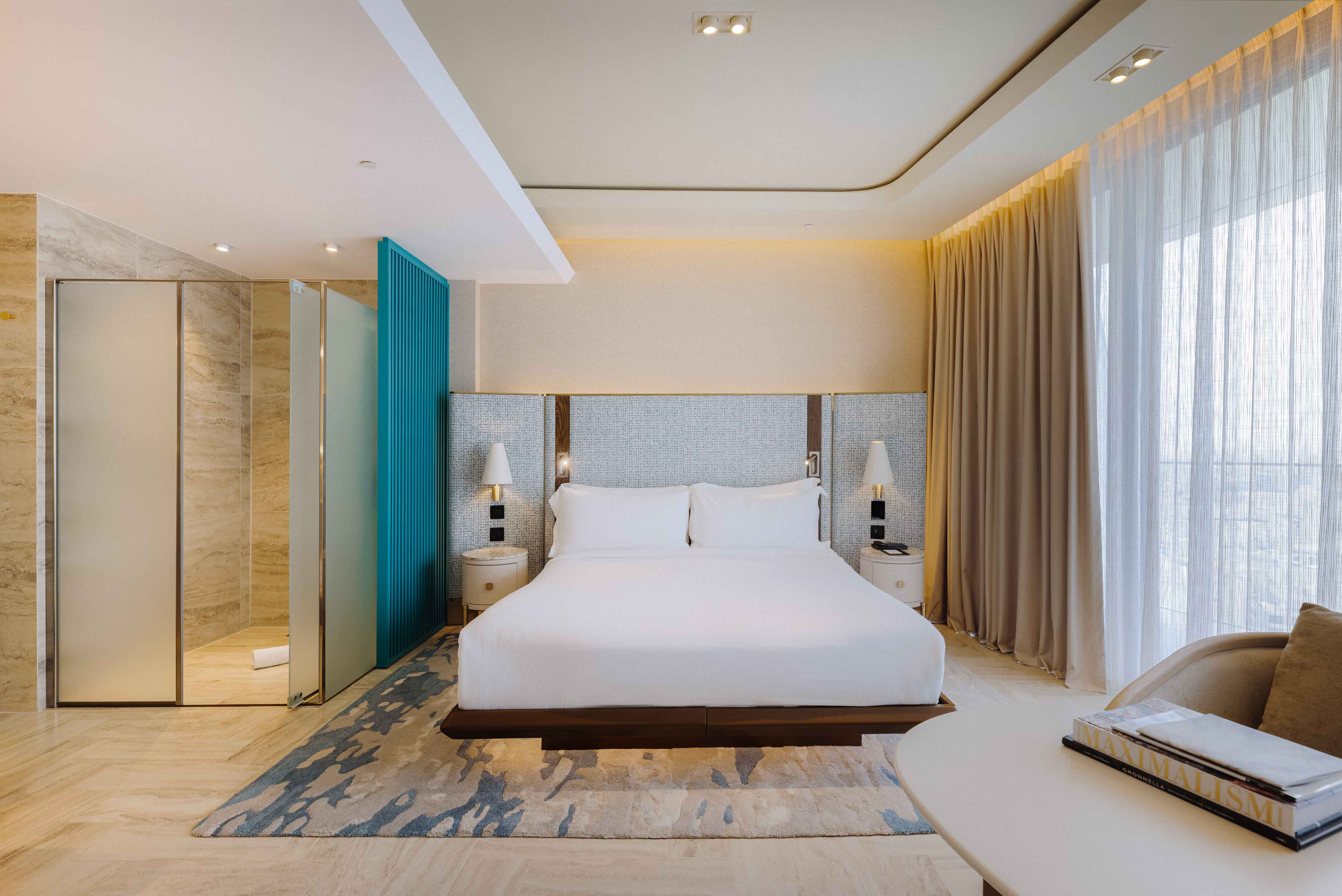 Modern hotel room with king bed, glass shower, teal accent, and sheer curtains.