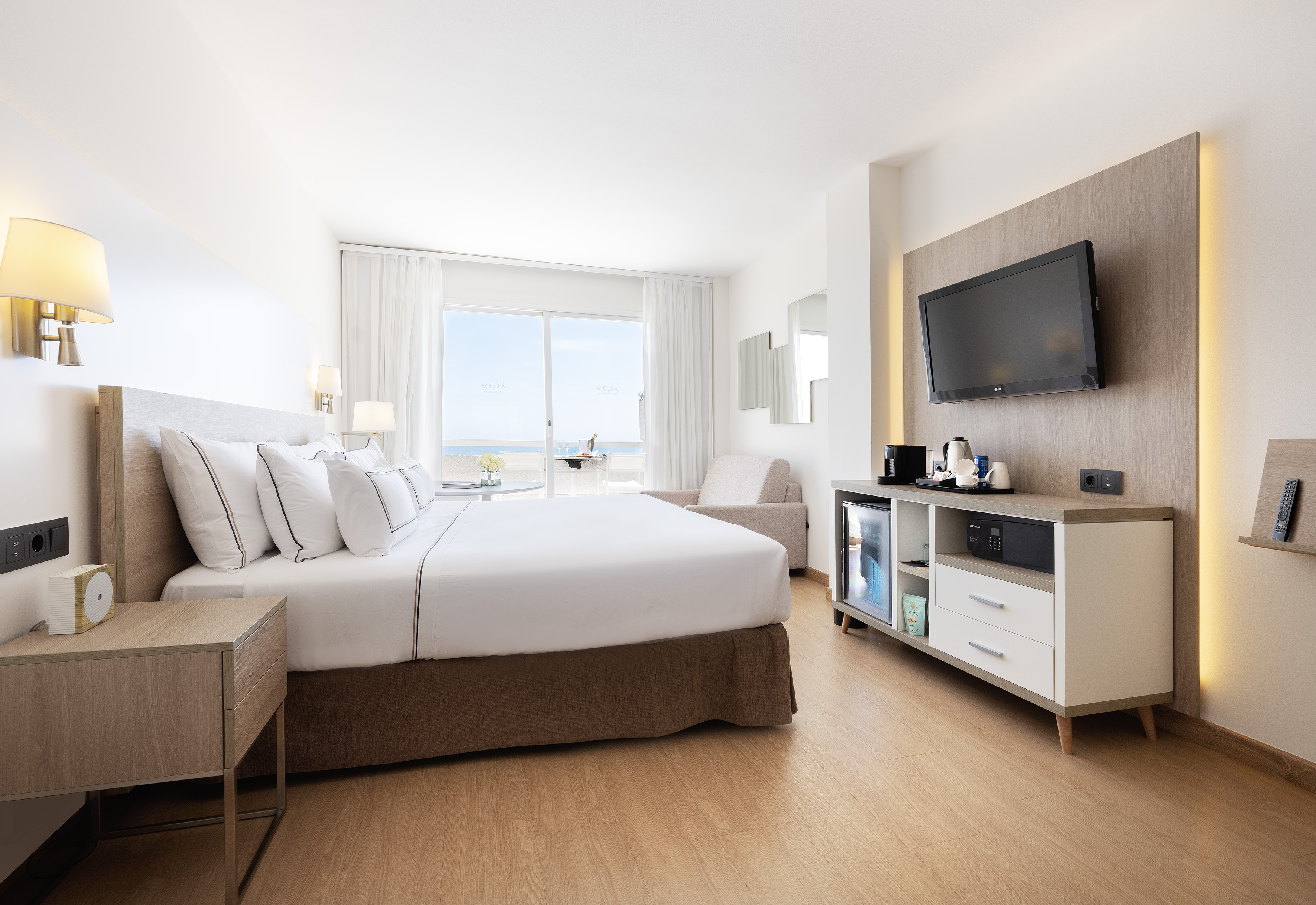 Modern hotel bedroom with ocean view balcony, large bed, TV, and minibar.