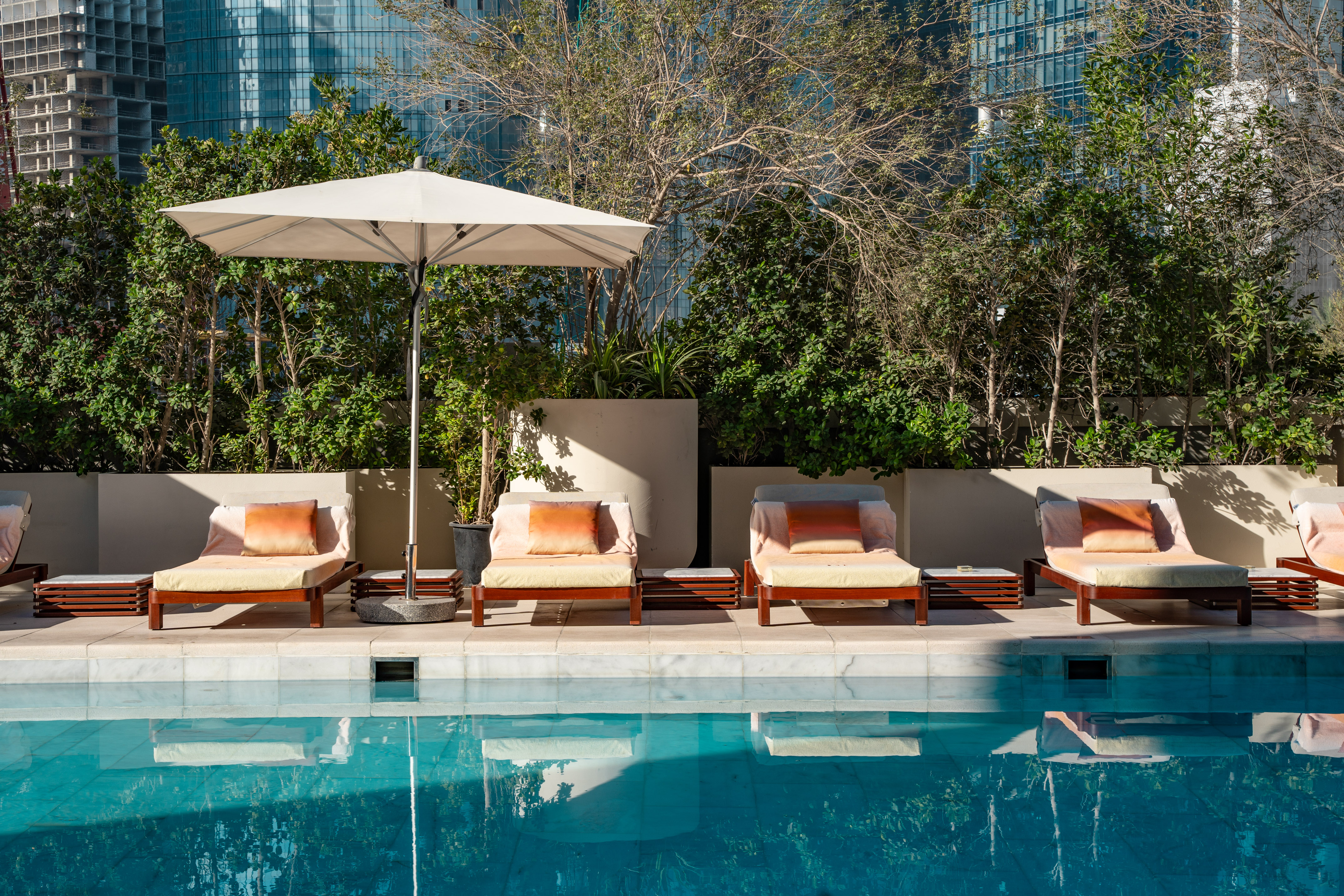 Outdoor pool with lounge chairs, umbrella, green foliage, and urban buildings.