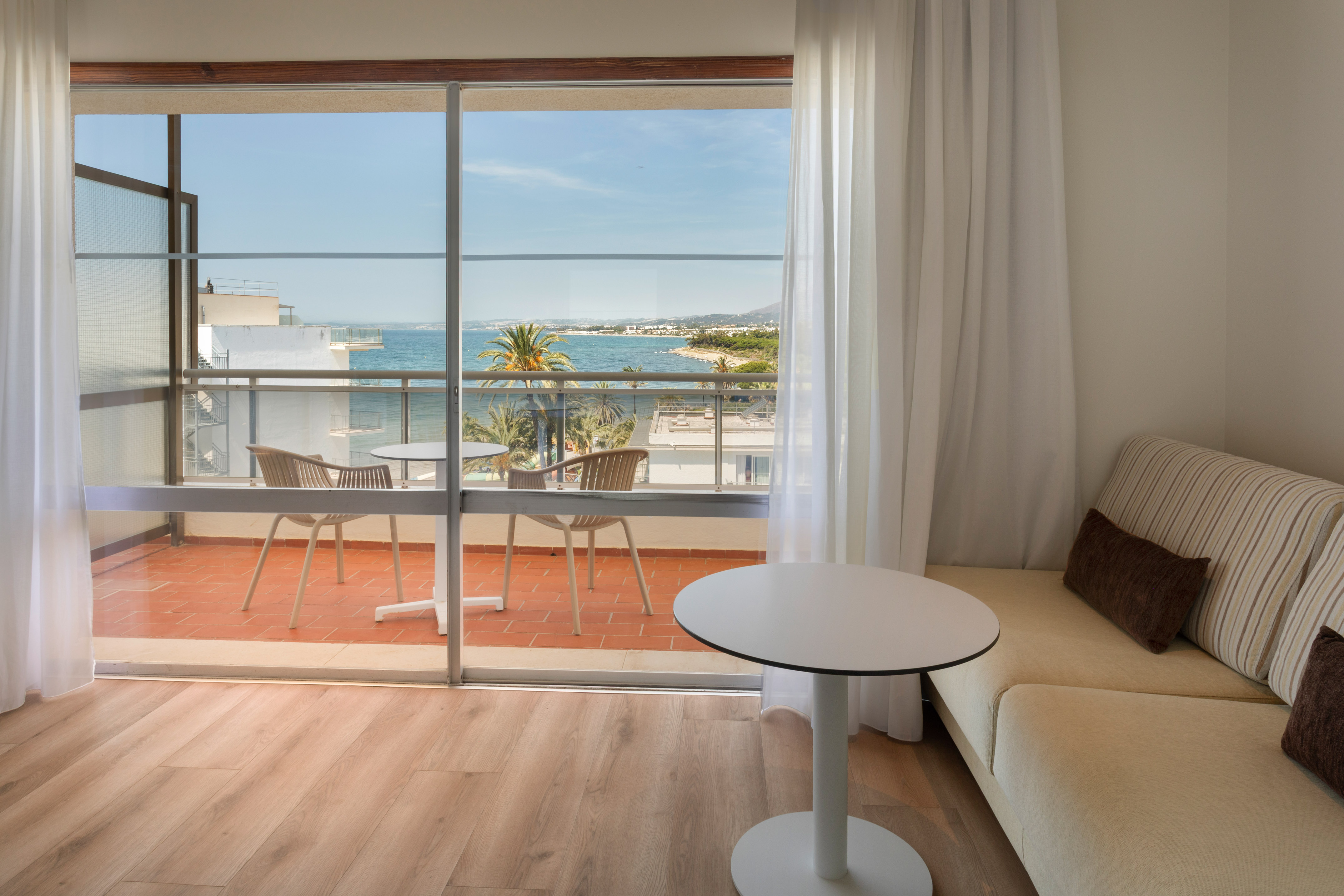 Bright hotel room with sea view balcony, palm trees, and coastal resort.