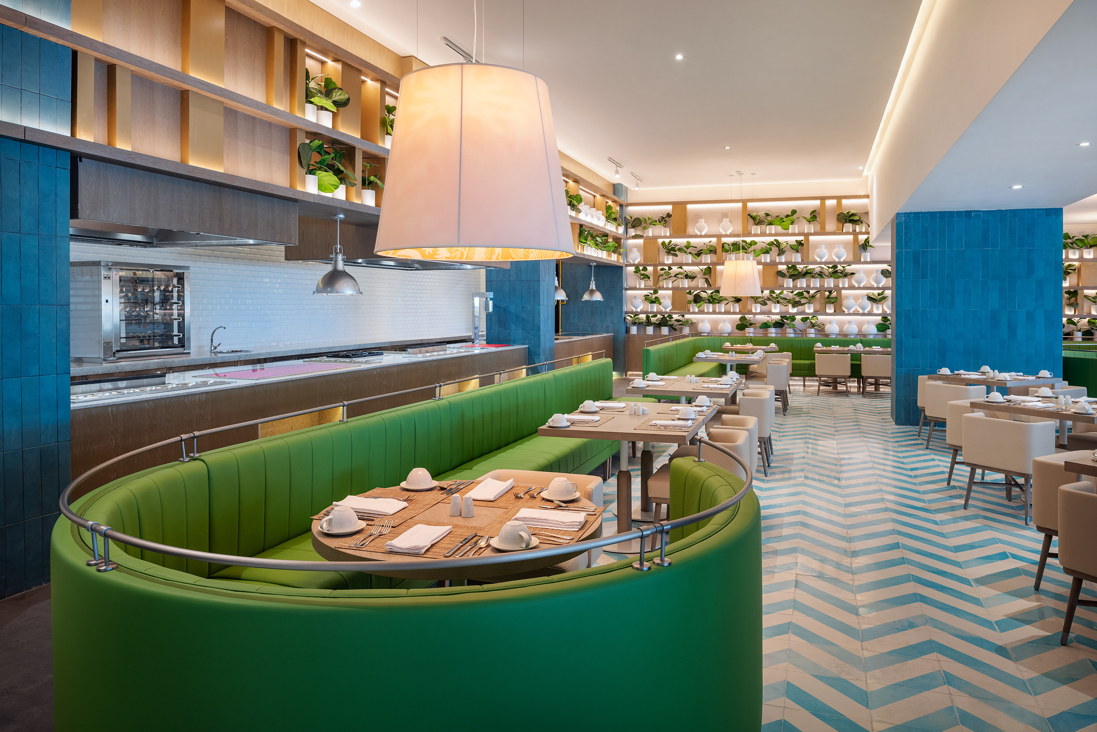 Modern restaurant with green booths, plant decor, buffet, and blue herringbone floor.